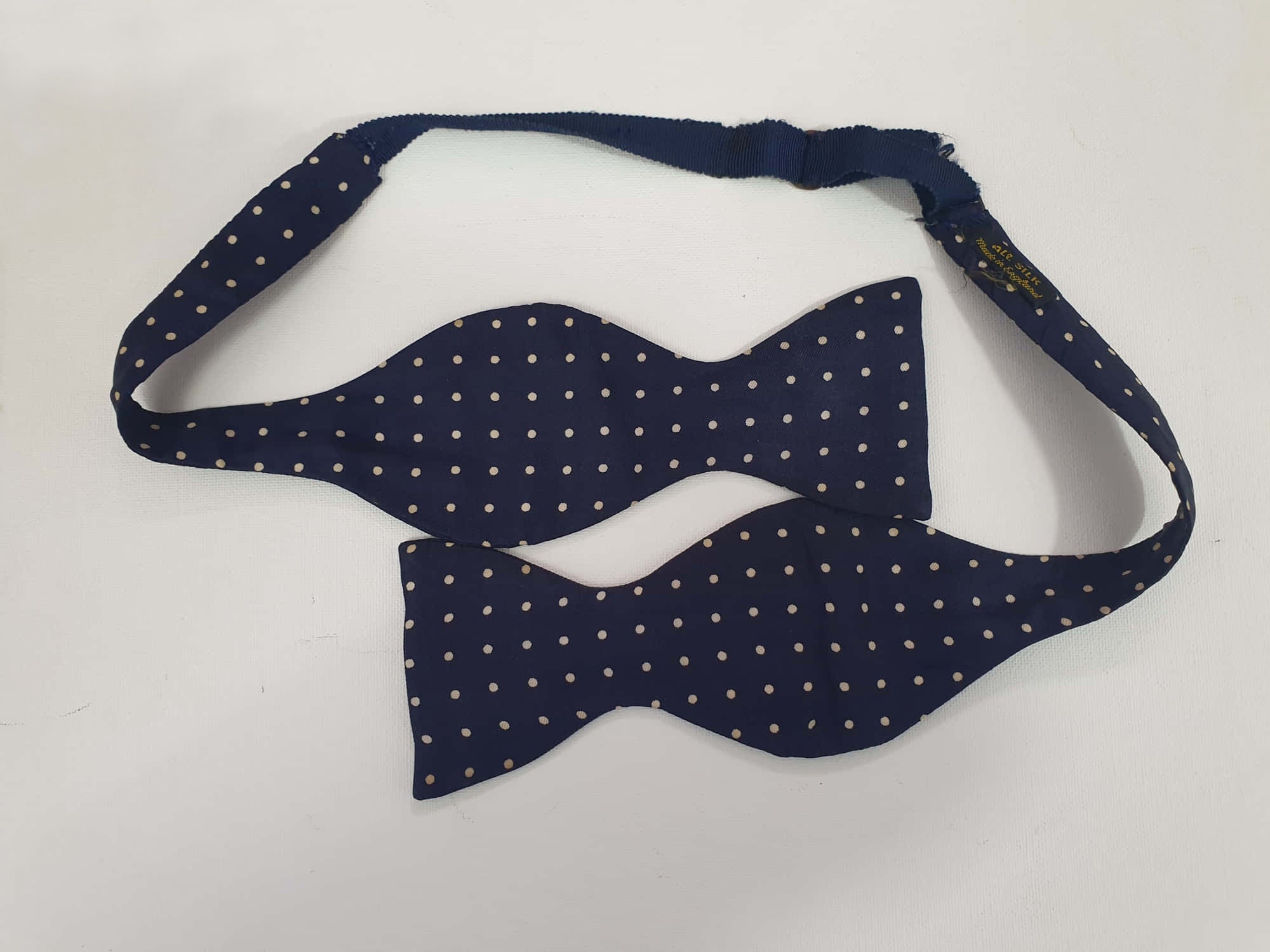 Navy blue polka dot self-tie bow tie has adjustable length. Ivory spots on navy blue ground.  Fabric is silk. Made in England - 1960s. Blade width 7.5 cm (3")