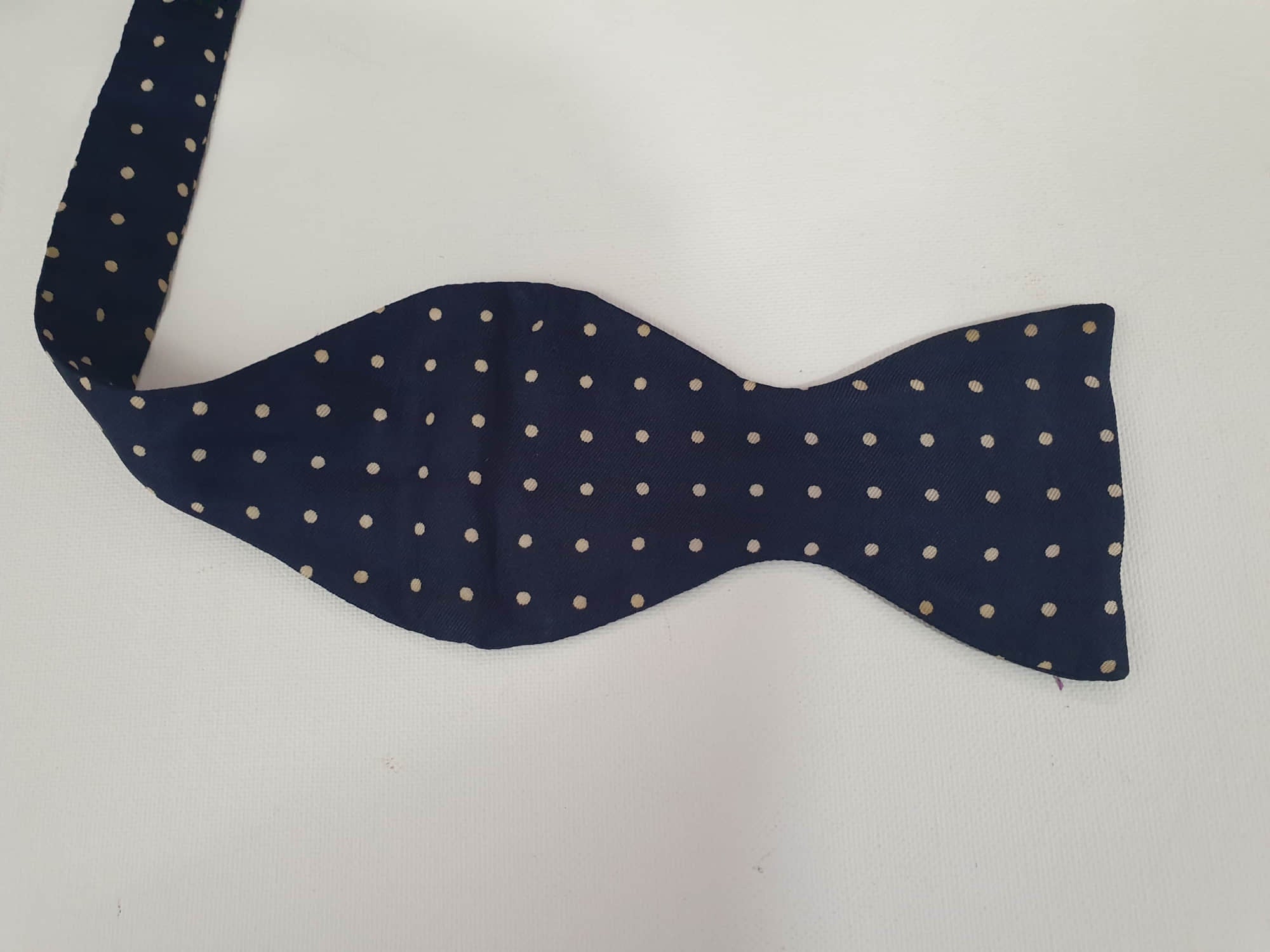 Navy blue polka dot self-tie bow tie has adjustable length. Ivory spots on navy blue ground.  Fabric is silk. Made in England - 1960s. Blade width 7.5 cm (3")