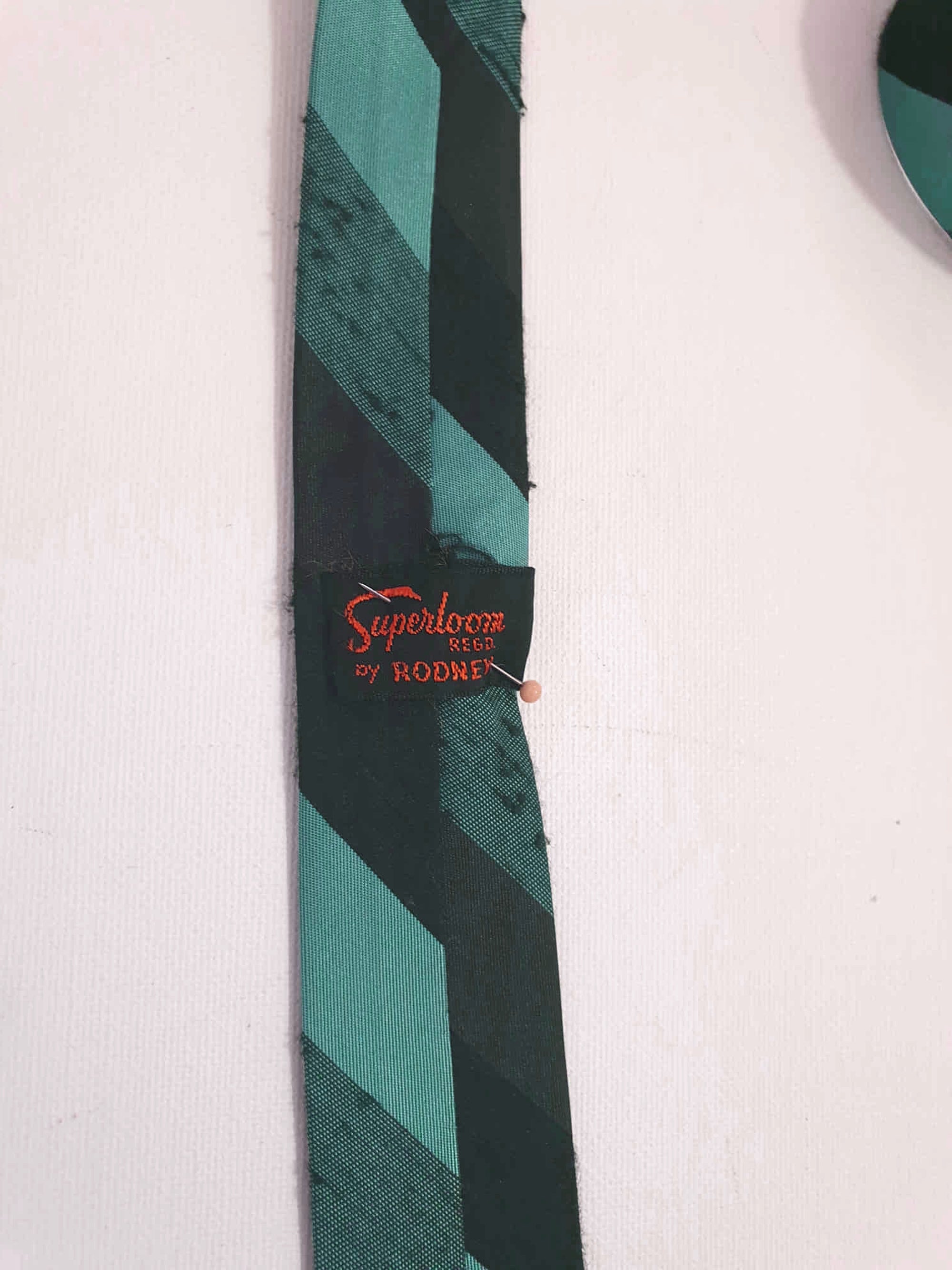 Super skinny tie in shades of grey-blue, grey and black. The grey band has a black fleck. Fabric is 100% polyester. Label reads "Superloom by Rodney" - 1960s. Blade width 3.7 cm (1.5") Length 130 cm (51") Excellent condition.