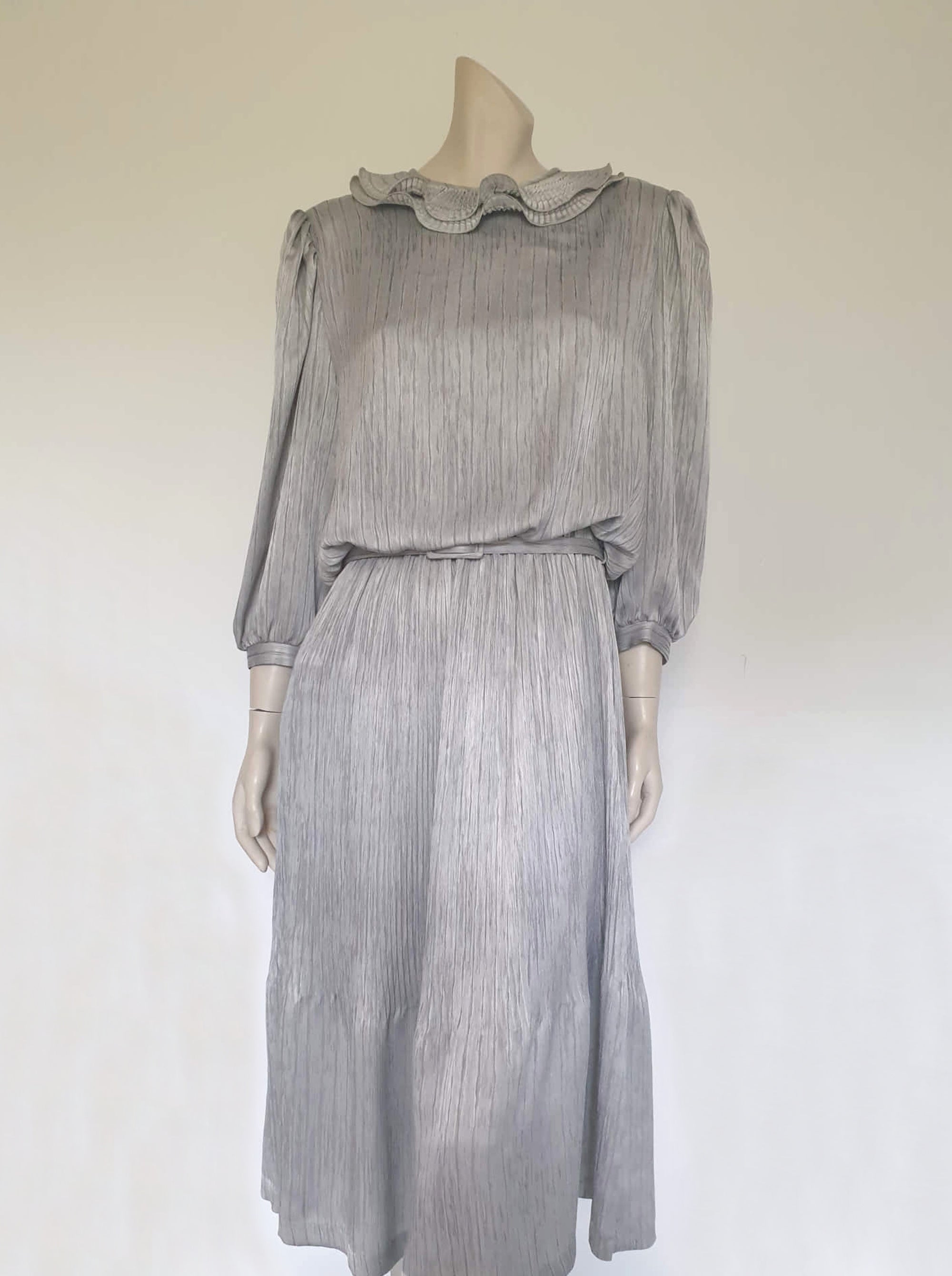 Beautiful and classy secretary style dress. Mid-calf length silver grey dress has three-quarter length buttoning sleeves and a circular ruffled collar. The skirt has very narrow permanent pleats to about knee level. The same pleated fabric has been used for the collar. The bodice is a full blouson look.  Brand is The Dress Company - 1980s. Medium