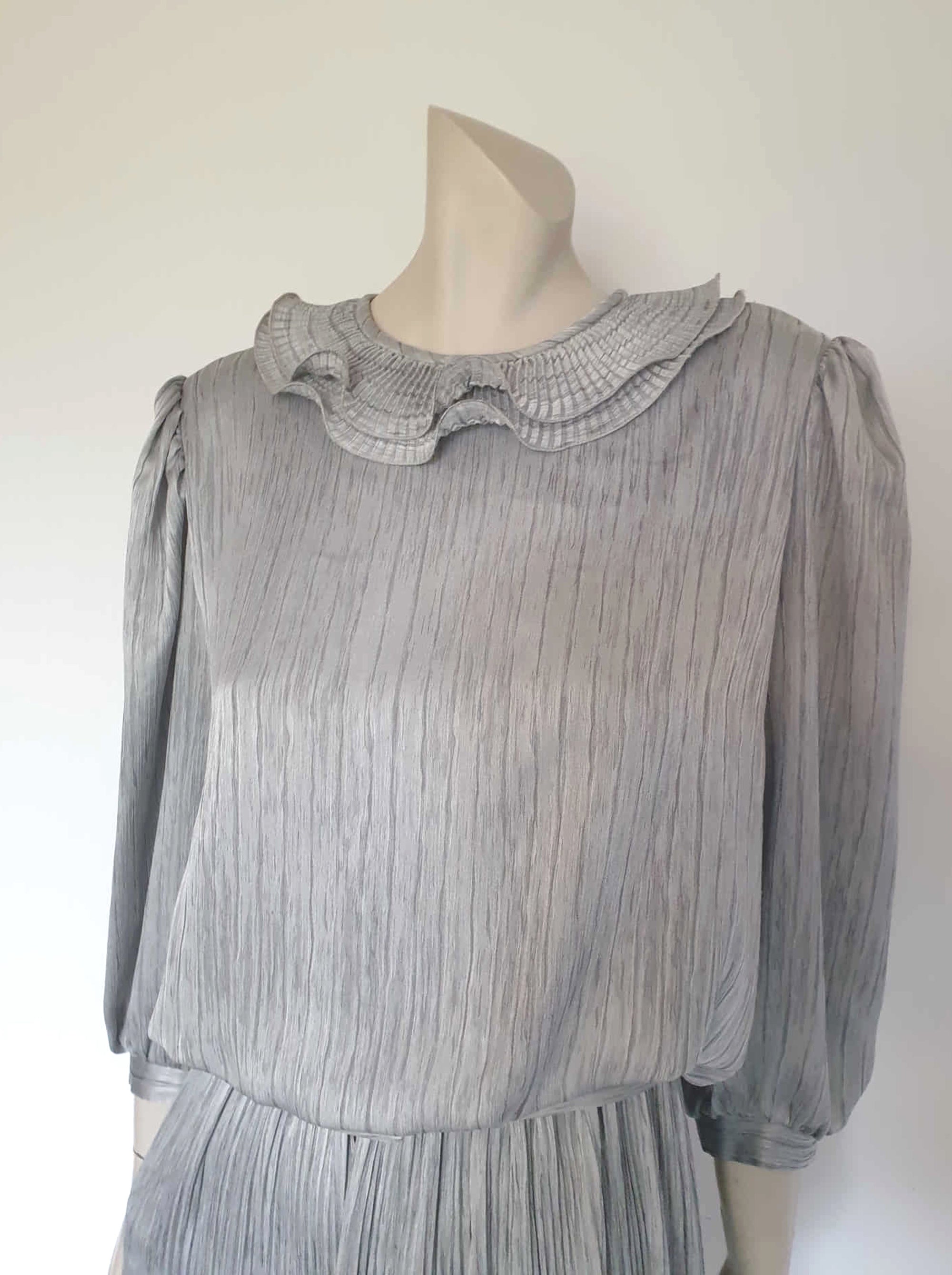 Beautiful and classy secretary style dress. Mid-calf length silver grey dress has three-quarter length buttoning sleeves and a circular ruffled collar. The skirt has very narrow permanent pleats to about knee level. The same pleated fabric has been used for the collar. The bodice is a full blouson look.  Brand is The Dress Company - 1980s. Medium