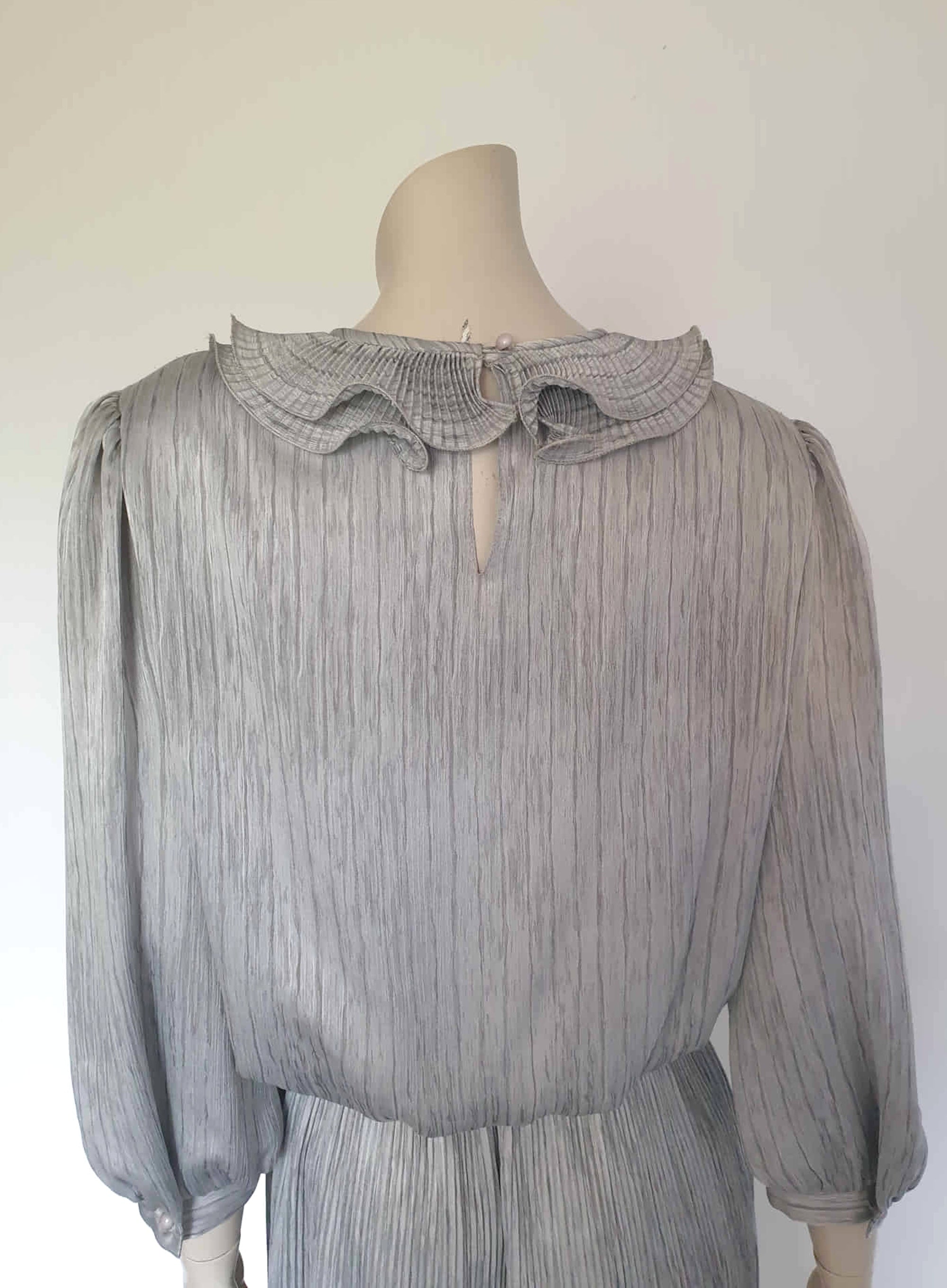 Beautiful and classy secretary style dress. Mid-calf length silver grey dress has three-quarter length buttoning sleeves and a circular ruffled collar. The skirt has very narrow permanent pleats to about knee level. The same pleated fabric has been used for the collar. The bodice is a full blouson look.  Brand is The Dress Company - 1980s. Medium