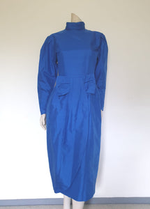 Screaming '80s winter formal dress from Argentinian born Sydney based designer Ewaldo Bock. Trimmed with an enormous bow at the front, this lower-calf  length dress features a high collar which closes at the back with three buttons and loops. The dress has long sleeves with dropped armholes pleated into the shoulder. The straight skirt has a shaped overlap at the front incorporated into the bow and a pleated rear vent. Closes with a zip at the back. The fabric is shantung silk.