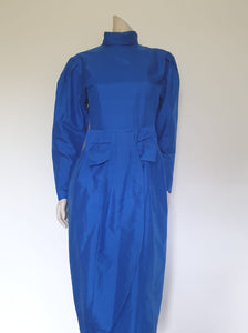 Screaming '80s winter formal dress from Argentinian born Sydney based designer Ewaldo Bock. Trimmed with an enormous bow at the front, this lower-calf  length dress features a high collar which closes at the back with three buttons and loops. The dress has long sleeves with dropped armholes pleated into the shoulder. The straight skirt has a shaped overlap at the front incorporated into the bow and a pleated rear vent. Closes with a zip at the back. The fabric is shantung silk.