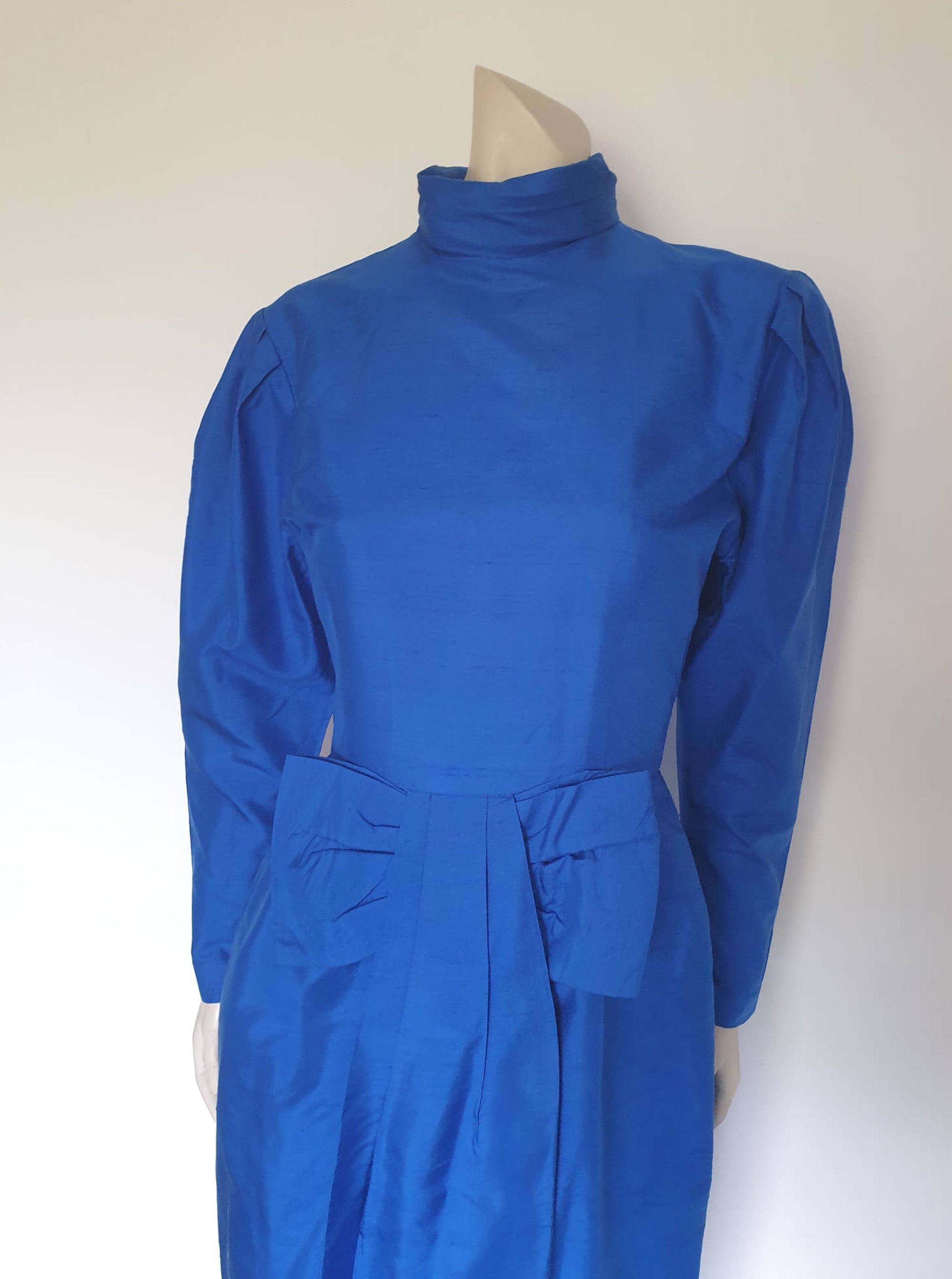 Screaming '80s winter formal dress from Argentinian born Sydney based designer Ewaldo Bock. Trimmed with an enormous bow at the front, this lower-calf  length dress features a high collar which closes at the back with three buttons and loops. The dress has long sleeves with dropped armholes pleated into the shoulder. The straight skirt has a shaped overlap at the front incorporated into the bow and a pleated rear vent. Closes with a zip at the back. The fabric is shantung silk.