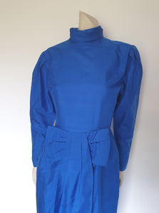 Screaming '80s winter formal dress from Argentinian born Sydney based designer Ewaldo Bock. Trimmed with an enormous bow at the front, this lower-calf  length dress features a high collar which closes at the back with three buttons and loops. The dress has long sleeves with dropped armholes pleated into the shoulder. The straight skirt has a shaped overlap at the front incorporated into the bow and a pleated rear vent. Closes with a zip at the back. The fabric is shantung silk.