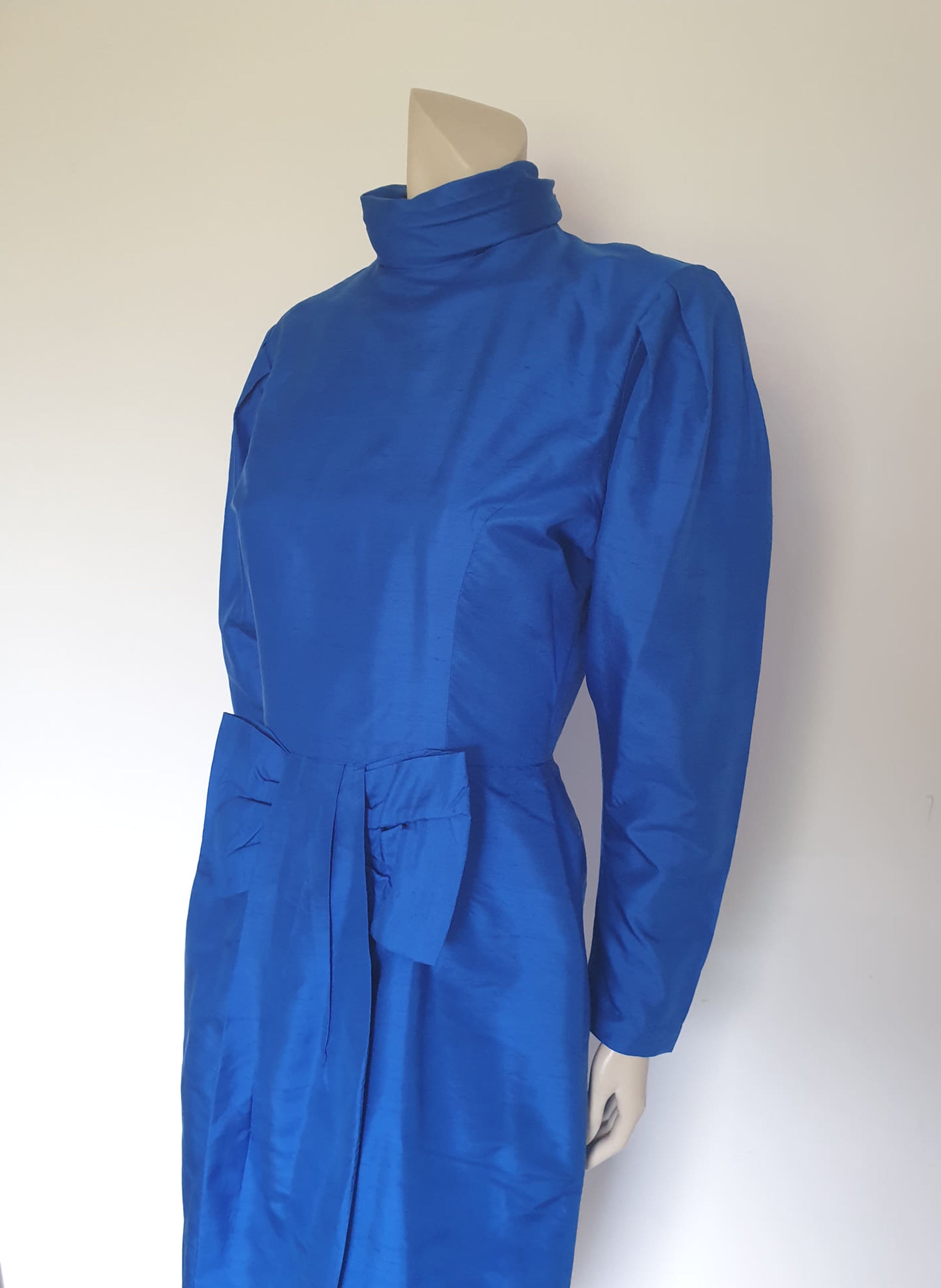 Screaming '80s winter formal dress from Argentinian born Sydney based designer Ewaldo Bock. Trimmed with an enormous bow at the front, this lower-calf  length dress features a high collar which closes at the back with three buttons and loops. The dress has long sleeves with dropped armholes pleated into the shoulder. The straight skirt has a shaped overlap at the front incorporated into the bow and a pleated rear vent. Closes with a zip at the back. The fabric is shantung silk.