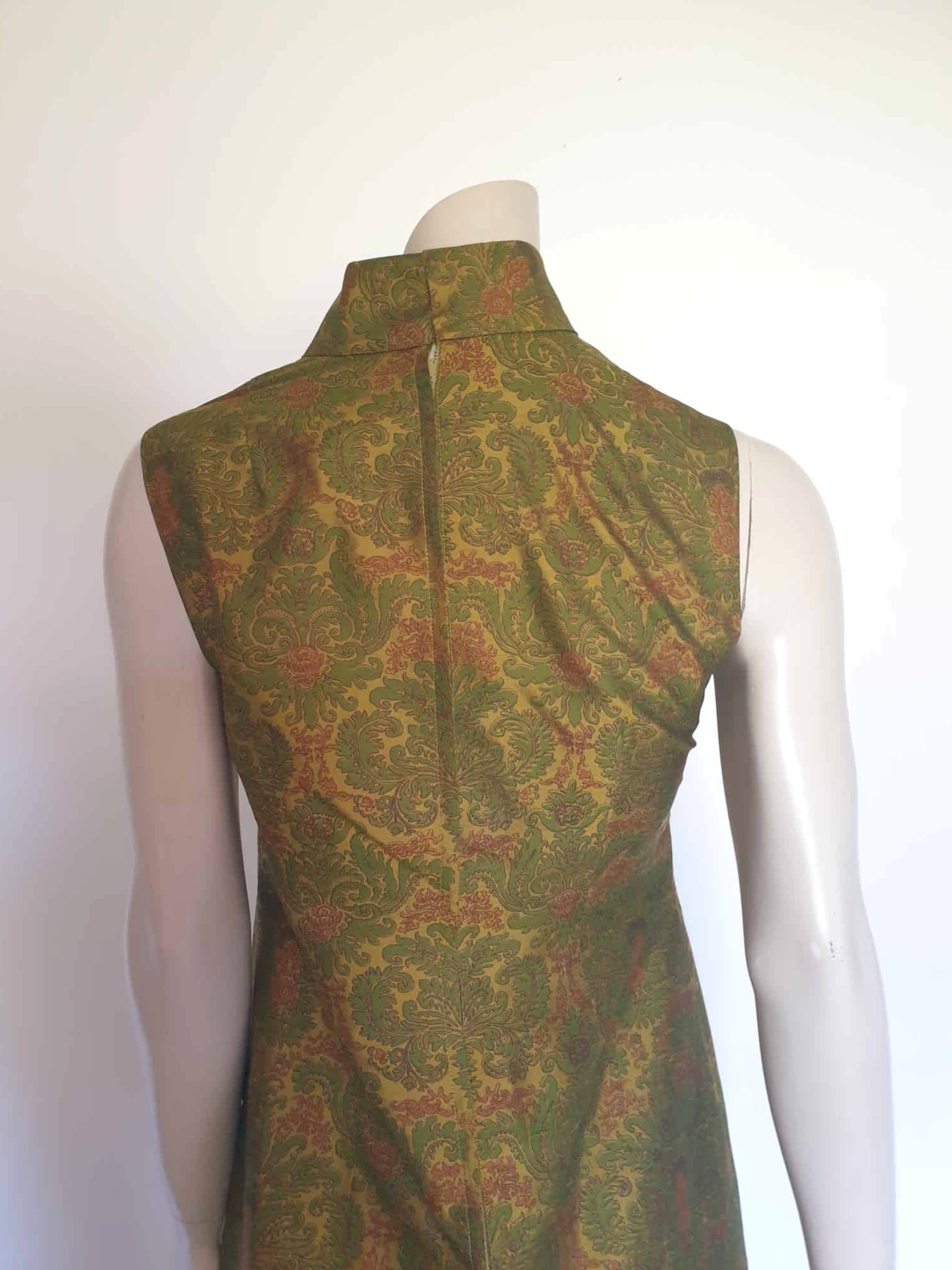 Gorgeous full length formal gown has a stylised Victorian style print in green and coral on a yellow-green ground and overall looks olive green. The fabric is stiff and has a sheen. It has a high standing collar. Closes at the back with a metal zip. The collar closes with hooks and eyes.  Fully lined. Custom made in Hong Kong by Jacky Taylor & Co. Late 1960s - early 1970s. Small.