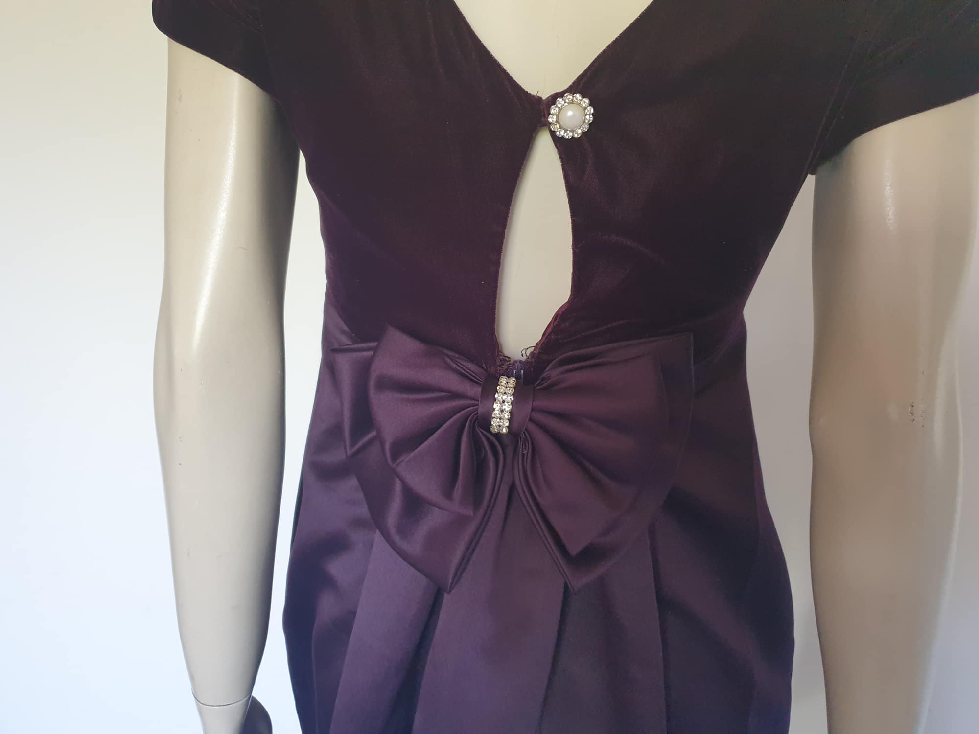 Beautiful and elegant aubergine formal sheath style gown with velvet bodice and stunning rear view. The gown has a wide neckline cut low at the back where it features a diamante studded bow extending into a narrow train over the the rear of the dress. Beneath the train the gown is slit to above the knee. The bodice is velvet and the satin skirt falls from beneath the bust. Closes at the back with a zip and a fancy button. Fabric is polyester. Made in Australia. Brand is Aurelie - 1980s. XS