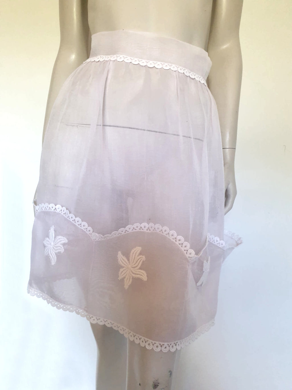 Hostess apron - decorative only. The half apron is palest pink sheer cotton organdy. Five ornamental scalloped pockets at the bottom feature white cotton floral motifs. White lacy cotton braid trims the edges as well as the waist. The straps are fixed with a little bow like ruffle. Hand made item - 1950s.