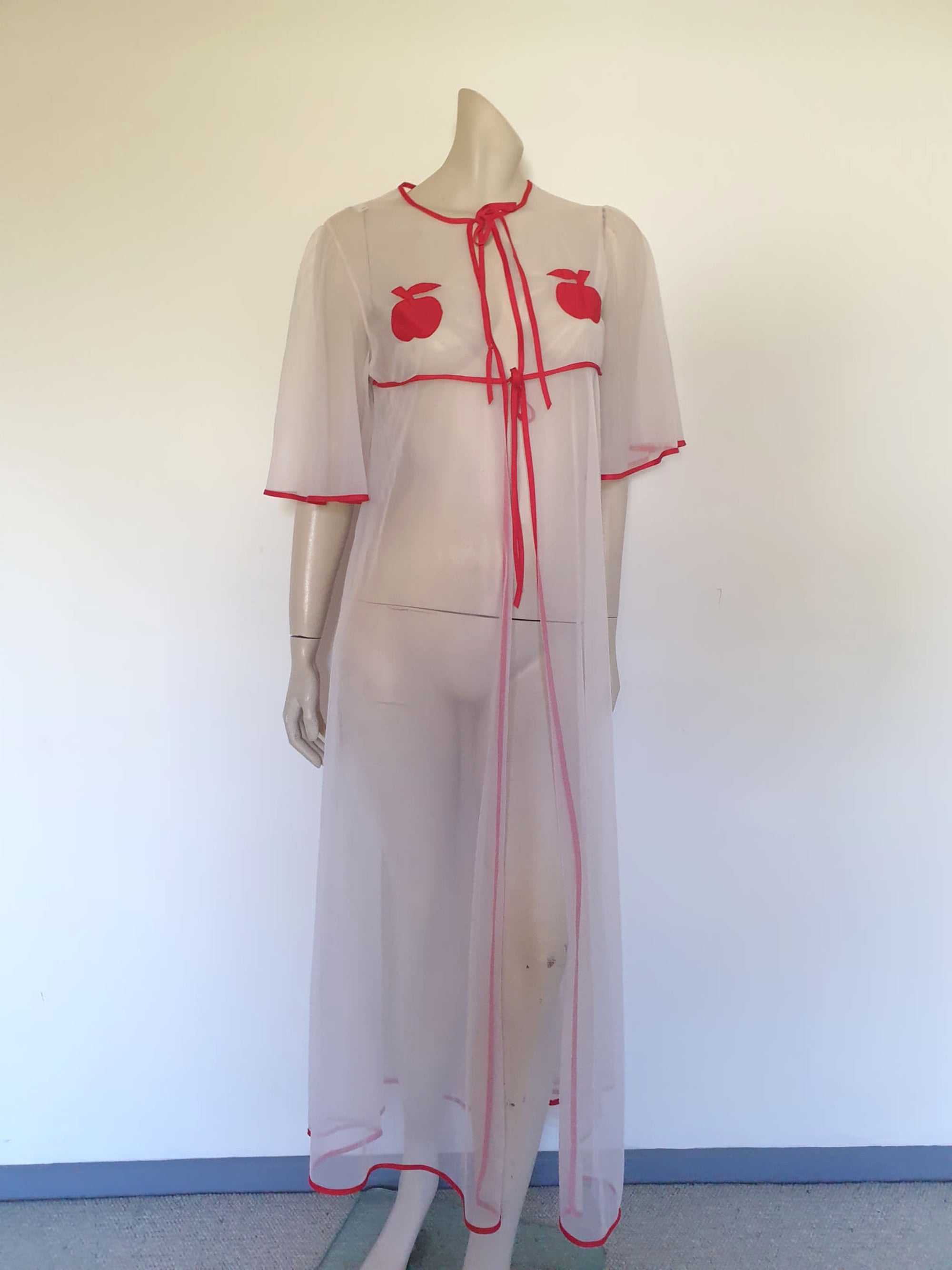 Super cute sheer white robe has red apples appliqued over the bust. The robe has full, elbow-length flutter sleeves and all edges are bound with red extending into ties at the neck and bust.   Ankle length. Fabric is polyamide (nylon). Made in Argentina. No brand label - 1970s. Medium