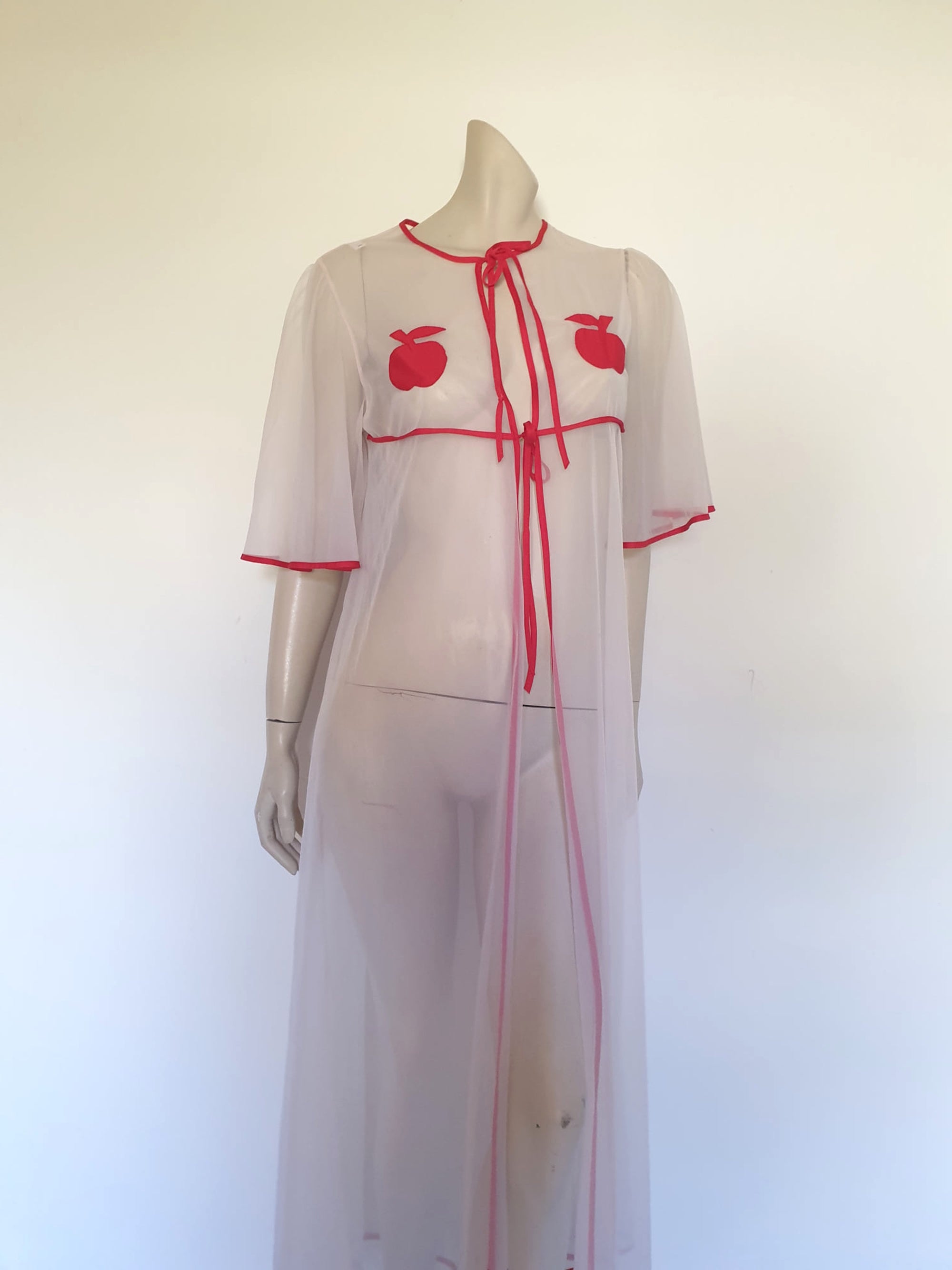 Super cute sheer white robe has red apples appliqued over the bust. The robe has full, elbow-length flutter sleeves and all edges are bound with red extending into ties at the neck and bust.   Ankle length. Fabric is polyamide (nylon). Made in Argentina. No brand label - 1970s. Medium