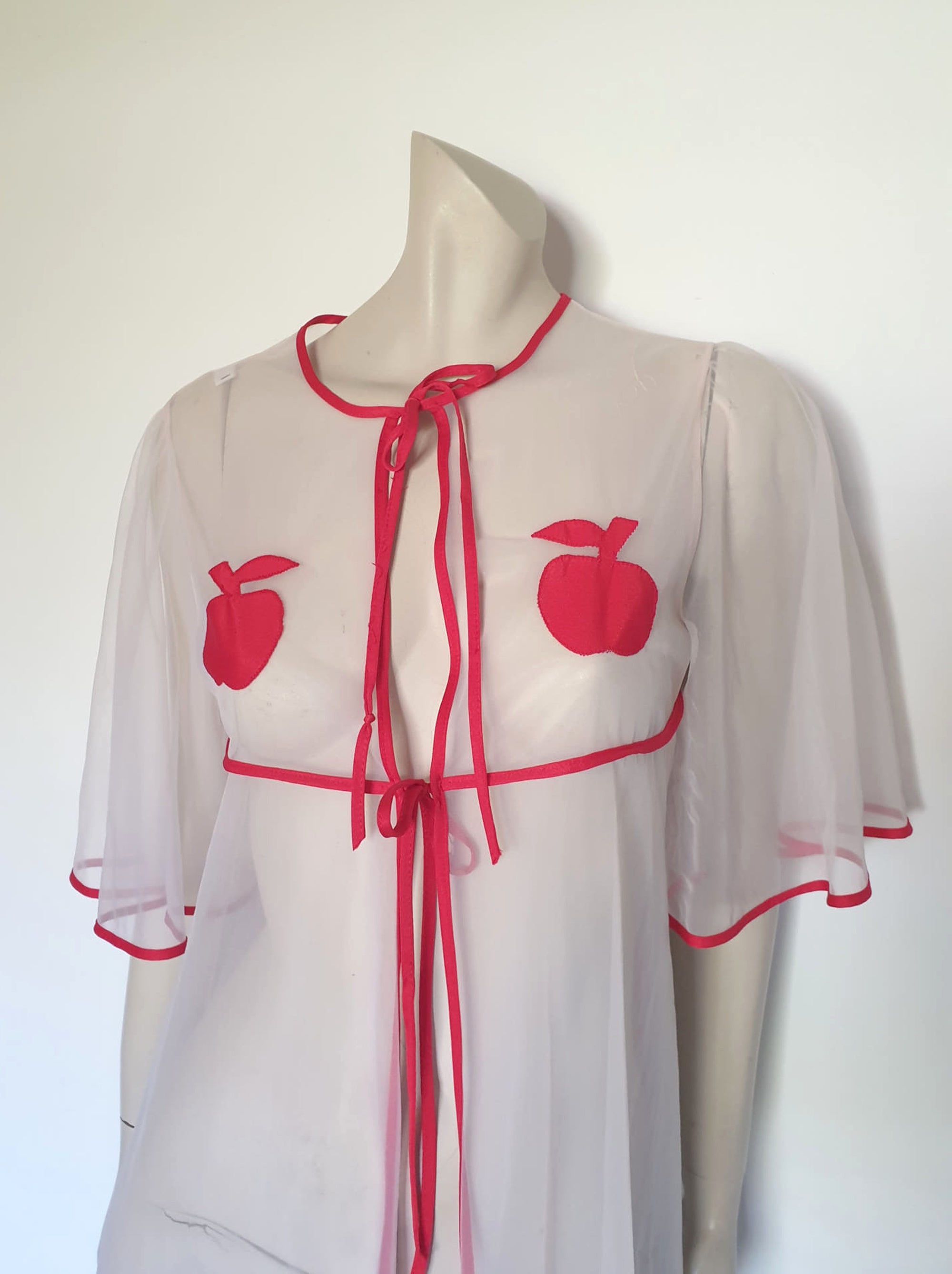 Super cute sheer white robe has red apples appliqued over the bust. The robe has full, elbow-length flutter sleeves and all edges are bound with red extending into ties at the neck and bust.   Ankle length. Fabric is polyamide (nylon). Made in Argentina. No brand label - 1970s. Medium