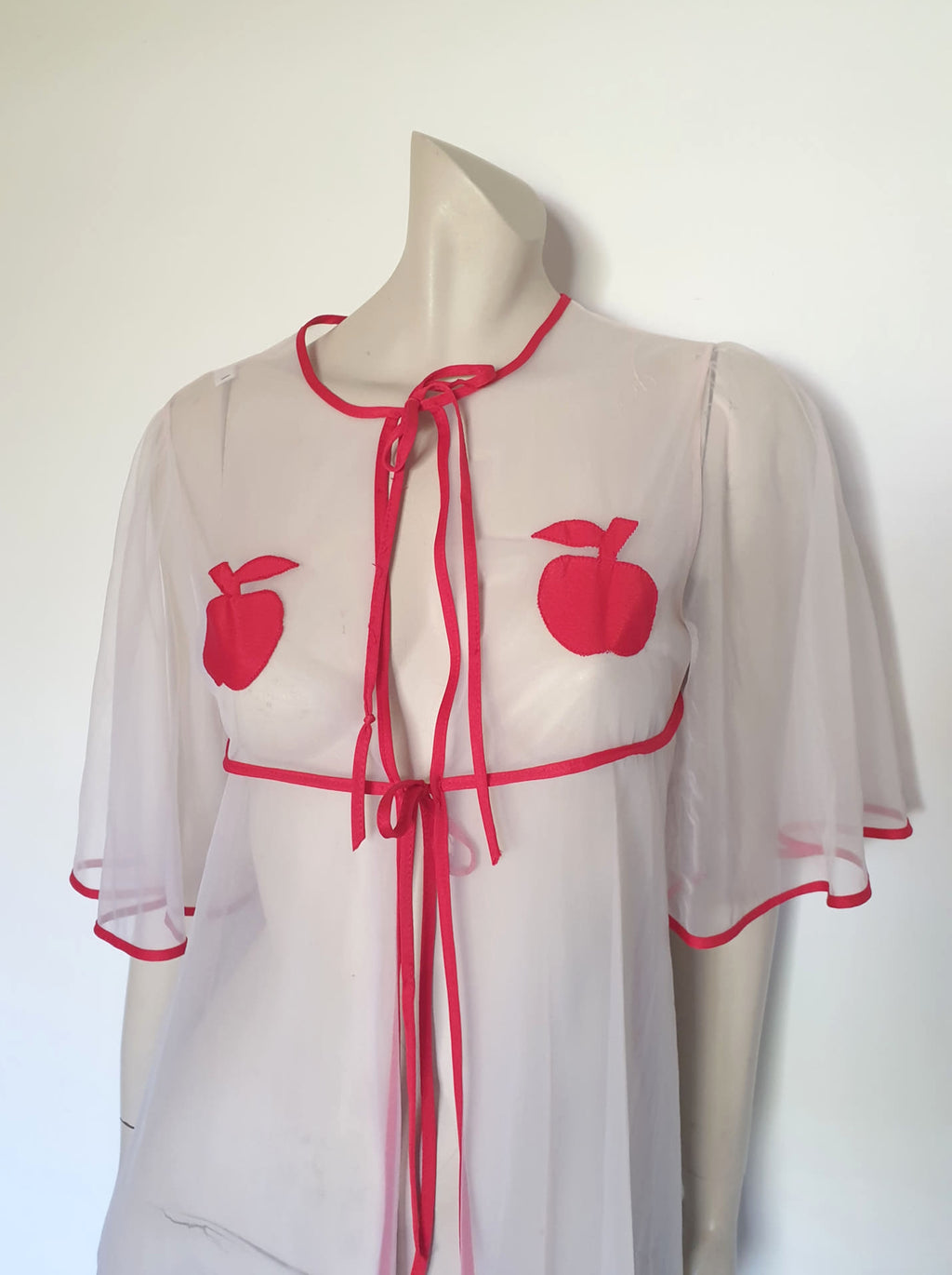 Super cute sheer white robe has red apples appliqued over the bust. The robe has full, elbow-length flutter sleeves and all edges are bound with red extending into ties at the neck and bust.   Ankle length. Fabric is polyamide (nylon). Made in Argentina. No brand label - 1970s. Medium