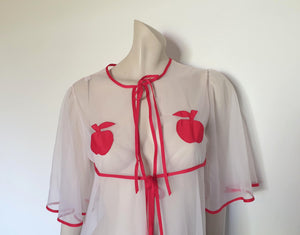 Super cute sheer white robe has red apples appliqued over the bust. The robe has full, elbow-length flutter sleeves and all edges are bound with red extending into ties at the neck and bust.   Ankle length. Fabric is polyamide (nylon). Made in Argentina. No brand label - 1970s. Medium