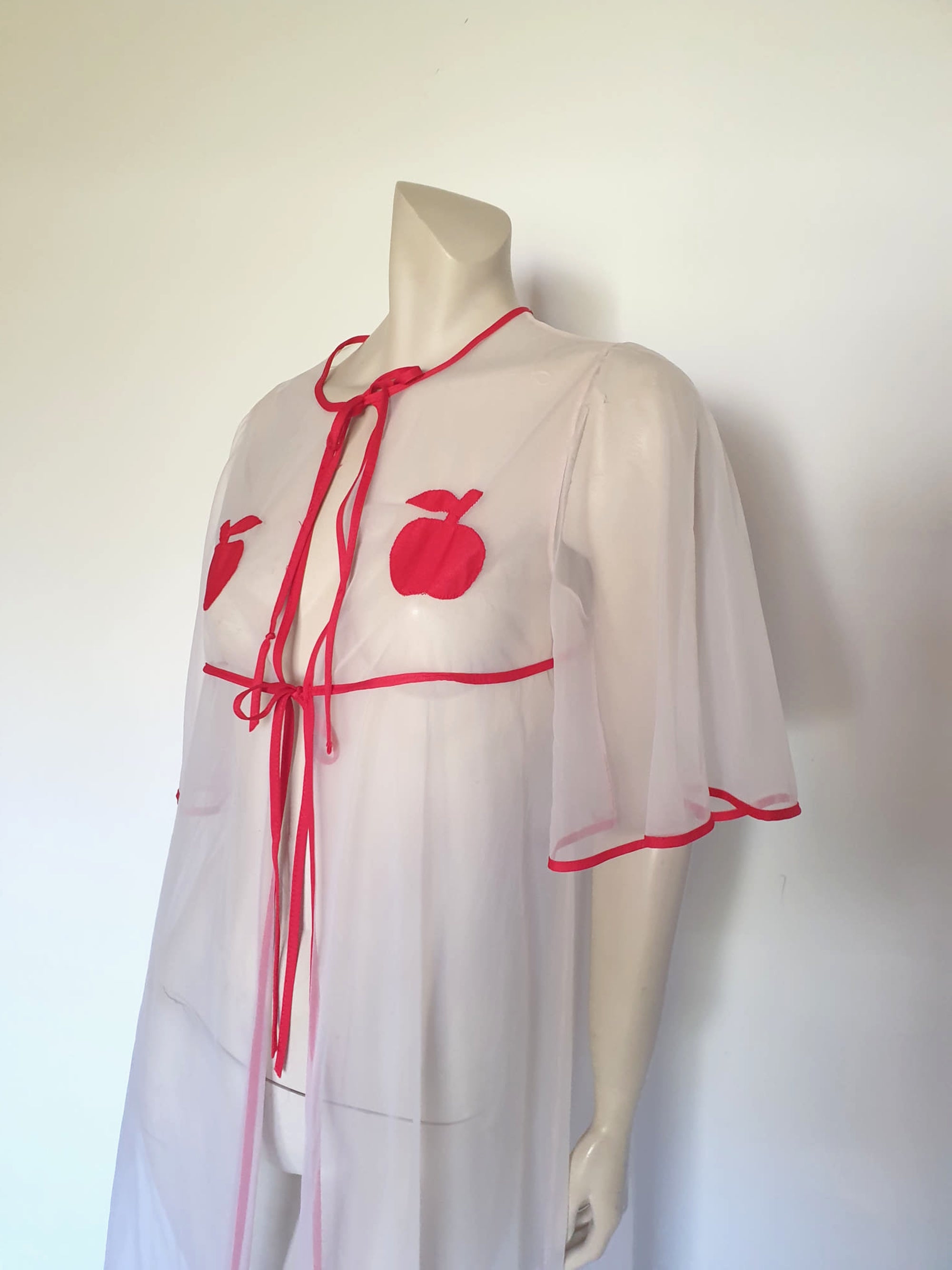 Super cute sheer white robe has red apples appliqued over the bust. The robe has full, elbow-length flutter sleeves and all edges are bound with red extending into ties at the neck and bust.   Ankle length. Fabric is polyamide (nylon). Made in Argentina. No brand label - 1970s. Medium