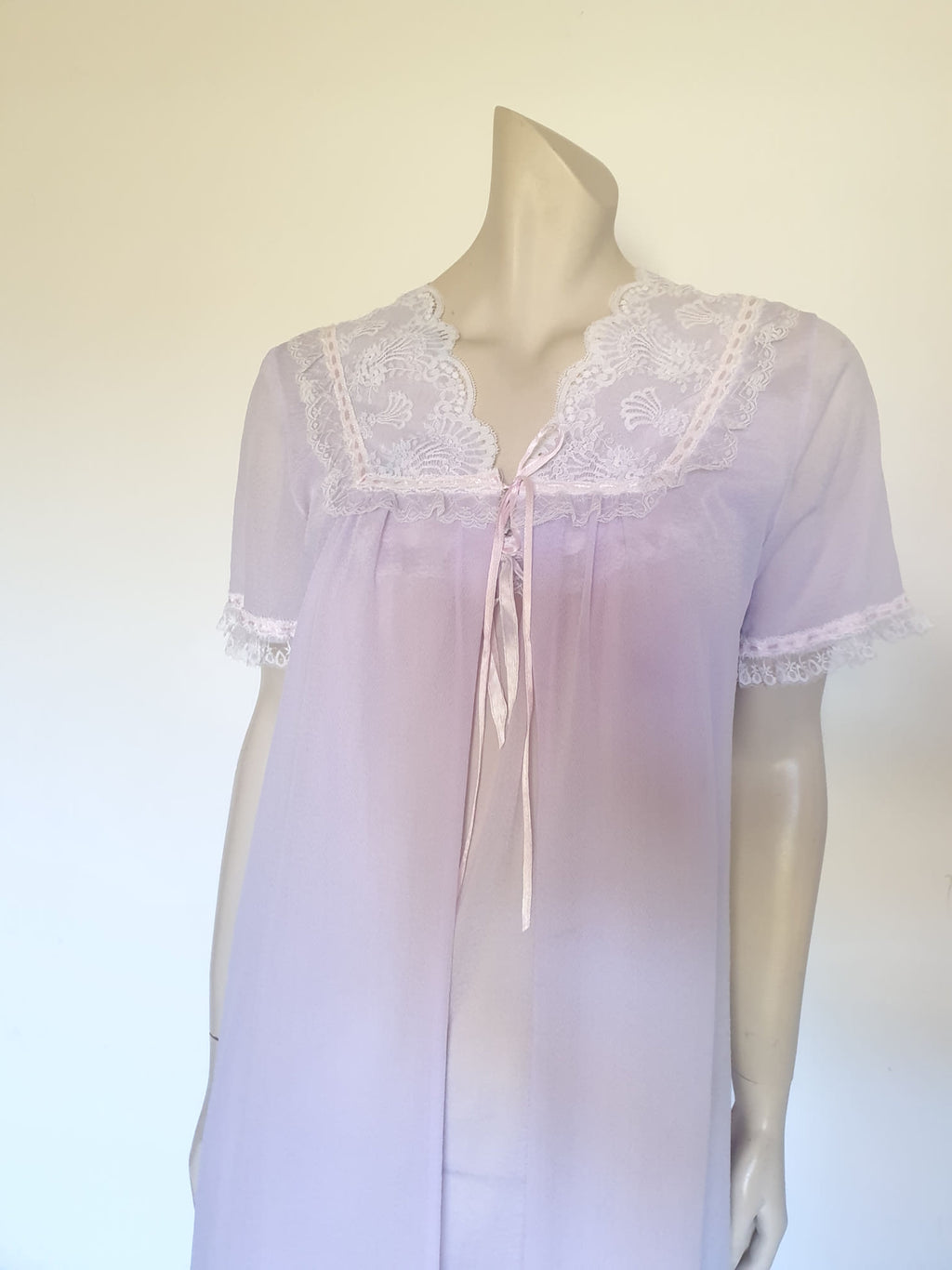 Pretty pale mauve crepe nightgown and robe set has a lace yoke tying at the neck with a ribbon on both the gown and robe. Pleas note: the colour is lighter than it appears in the photos. Above-knee length set has a high V neck and lace and pink ribbon trim at the yoke and sleeves. Hemline is also lace trimmed.  The robe has short sleeves, the gown is sleeveless. Fabric is nylon crepe. Brand is Irene - 1970s. Medium.