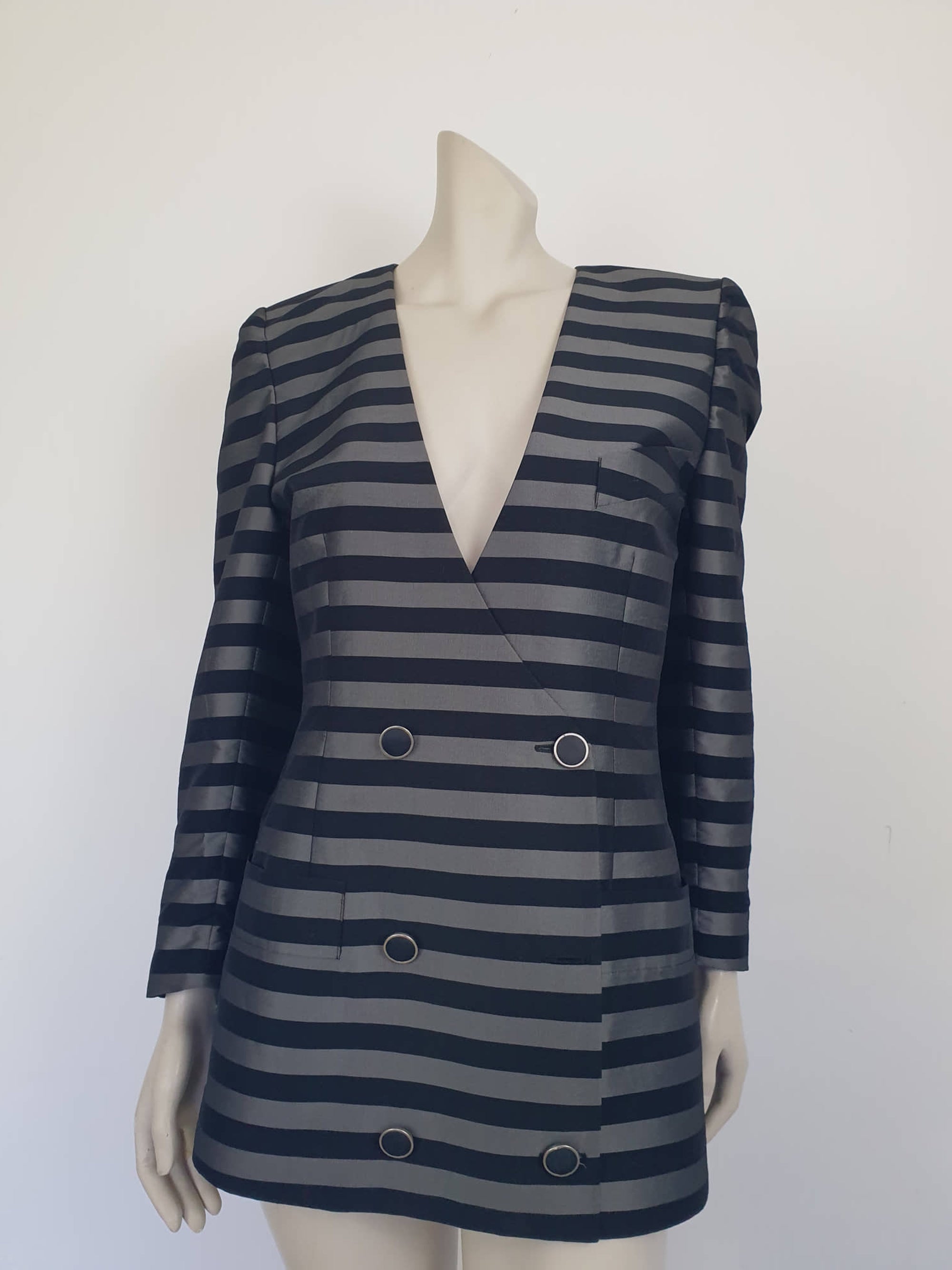 Grey and black striped longline jacket by Australian designer Teena Varigos. 
This V-necked jacket is truly double-breasted and can be worn buttoned either left over right or right over left. It's below hip length. It has two functional front pockets and a breast pocket. Shoulders are emphasised in true '80s style. The sleeves taper to the wrist. Two button sleeve trim and six button front stance. Fabric has the look of silk but is actually polyester - no fabric labels. Teena Varigos - 1980s.