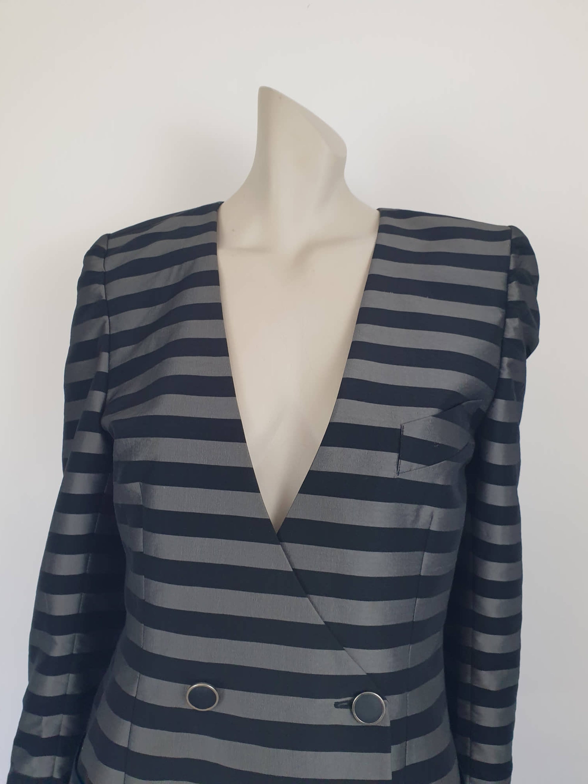 Grey and black striped longline jacket by Australian designer Teena Varigos. 
This V-necked jacket is truly double-breasted and can be worn buttoned either left over right or right over left. It's below hip length. It has two functional front pockets and a breast pocket. Shoulders are emphasised in true '80s style. The sleeves taper to the wrist. Two button sleeve trim and six button front stance. Fabric has the look of silk but is actually polyester - no fabric labels. Teena Varigos - 1980s.