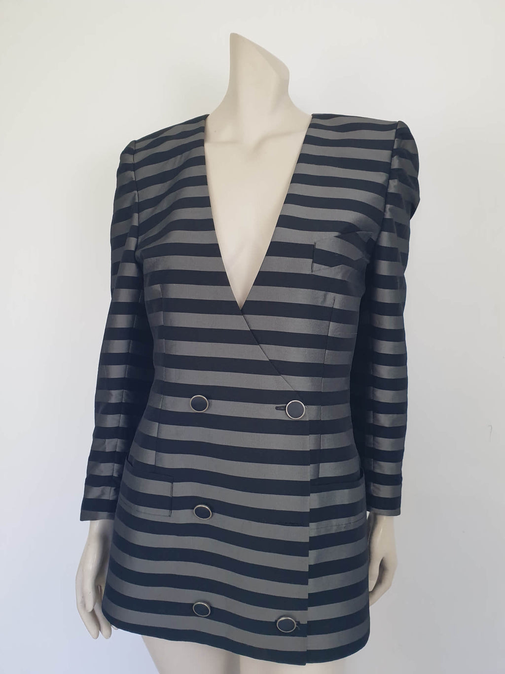 Grey and black striped longline jacket by Australian designer Teena Varigos. 
This V-necked jacket is truly double-breasted and can be worn buttoned either left over right or right over left. It's below hip length. It has two functional front pockets and a breast pocket. Shoulders are emphasised in true '80s style. The sleeves taper to the wrist. Two button sleeve trim and six button front stance. Fabric has the look of silk but is actually polyester - no fabric labels. Teena Varigos - 1980s.