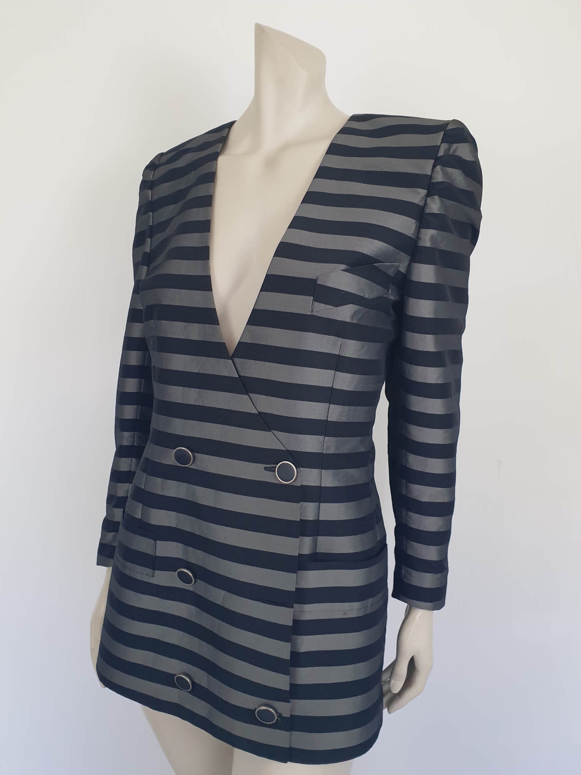 Grey and black striped longline jacket by Australian designer Teena Varigos. 
This V-necked jacket is truly double-breasted and can be worn buttoned either left over right or right over left. It's below hip length. It has two functional front pockets and a breast pocket. Shoulders are emphasised in true '80s style. The sleeves taper to the wrist. Two button sleeve trim and six button front stance. Fabric has the look of silk but is actually polyester - no fabric labels. Teena Varigos - 1980s.