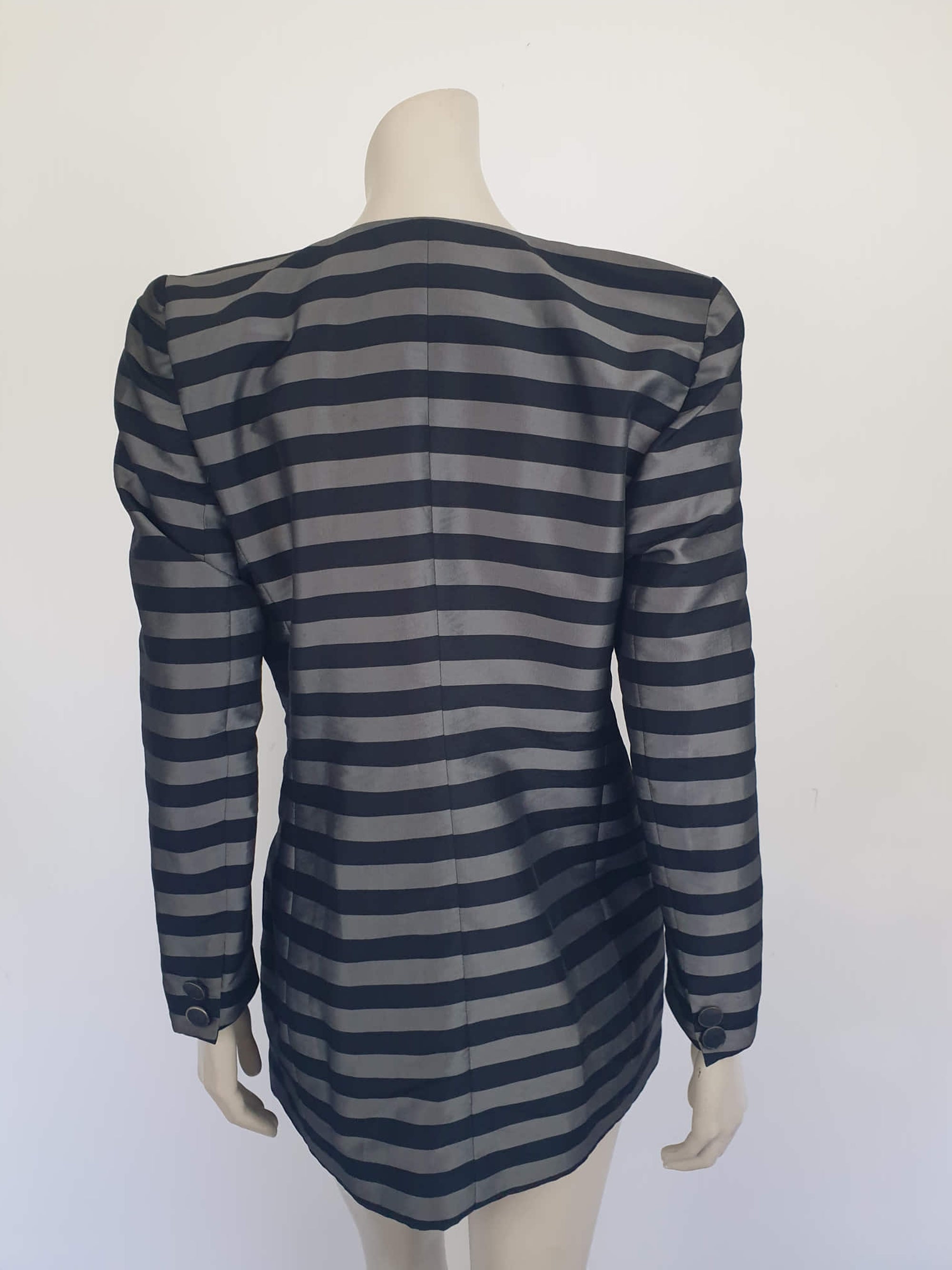 Grey and black striped longline jacket by Australian designer Teena Varigos. 
This V-necked jacket is truly double-breasted and can be worn buttoned either left over right or right over left. It's below hip length. It has two functional front pockets and a breast pocket. Shoulders are emphasised in true '80s style. The sleeves taper to the wrist. Two button sleeve trim and six button front stance. Fabric has the look of silk but is actually polyester - no fabric labels. Teena Varigos - 1980s.