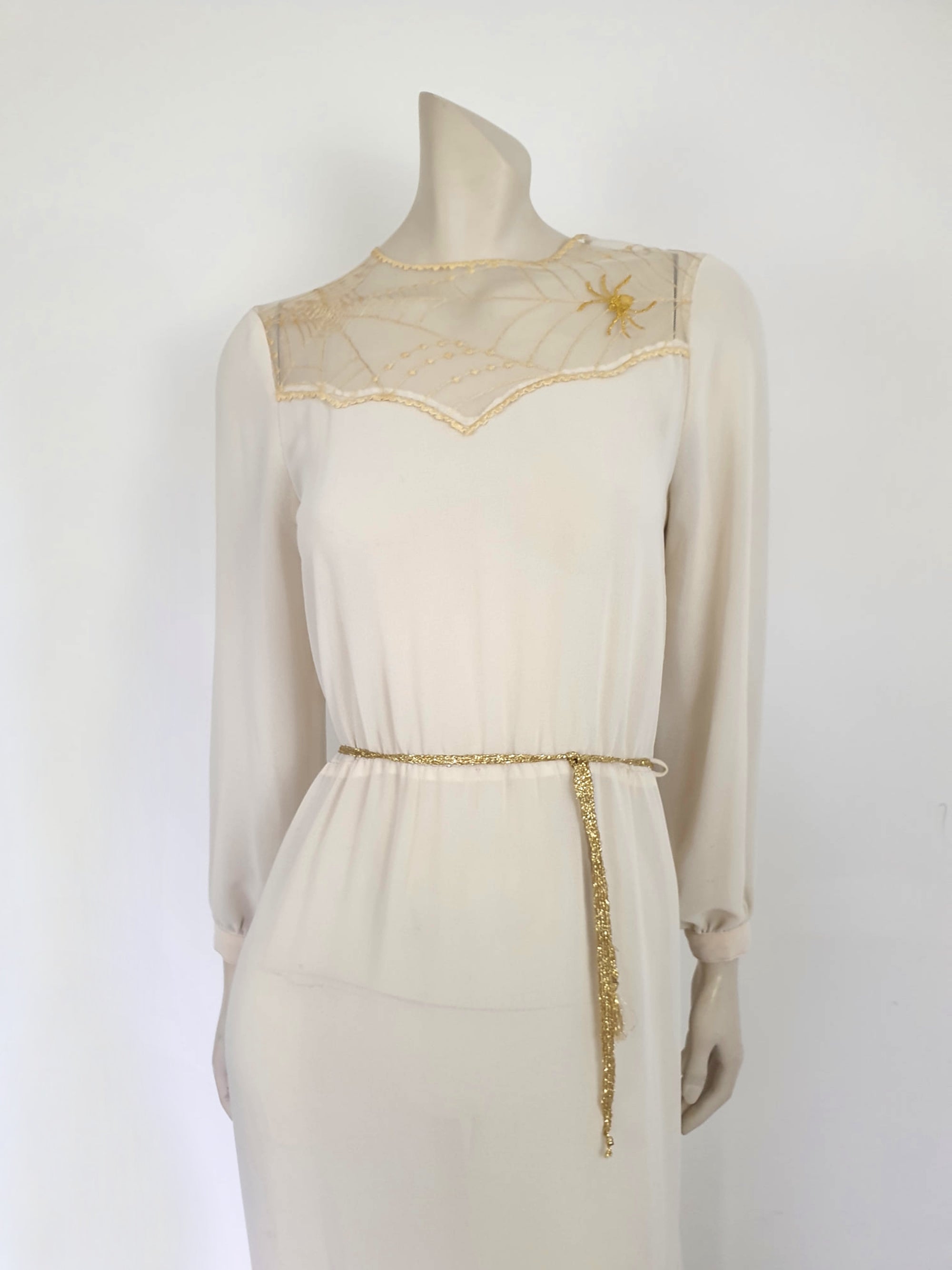 The perfect dress for Halloween, or for those who love spiders and spooky all year round. Pale beige (almost cream) crepe dress with an amazing yoke embroidered with a spiderweb, complete with gold spider. Dress is below-knee length, and has long sleeves which button at the cuffs. It just pulls on over the head and has one button and loop closure at the back neck. There is a separate gold tinsel tie for the waist which runs through carriers. Fabric is polyester. Brand is Ojay - 1970s. Medium.