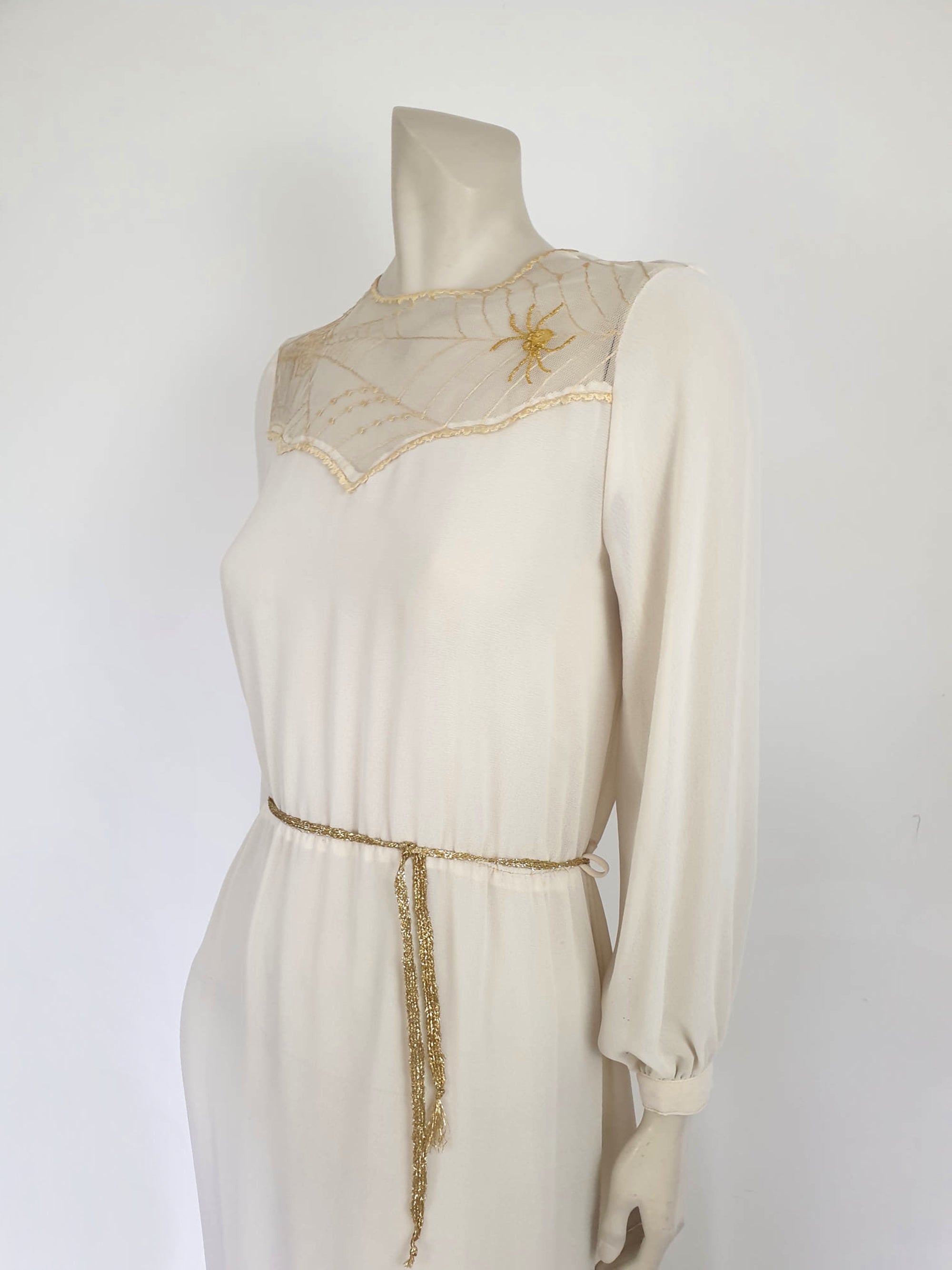 The perfect dress for Halloween, or for those who love spiders and spooky all year round. Pale beige (almost cream) crepe dress with an amazing yoke embroidered with a spiderweb, complete with gold spider. Dress is below-knee length, and has long sleeves which button at the cuffs. It just pulls on over the head and has one button and loop closure at the back neck. There is a separate gold tinsel tie for the waist which runs through carriers. Fabric is polyester. Brand is Ojay - 1970s. Medium.