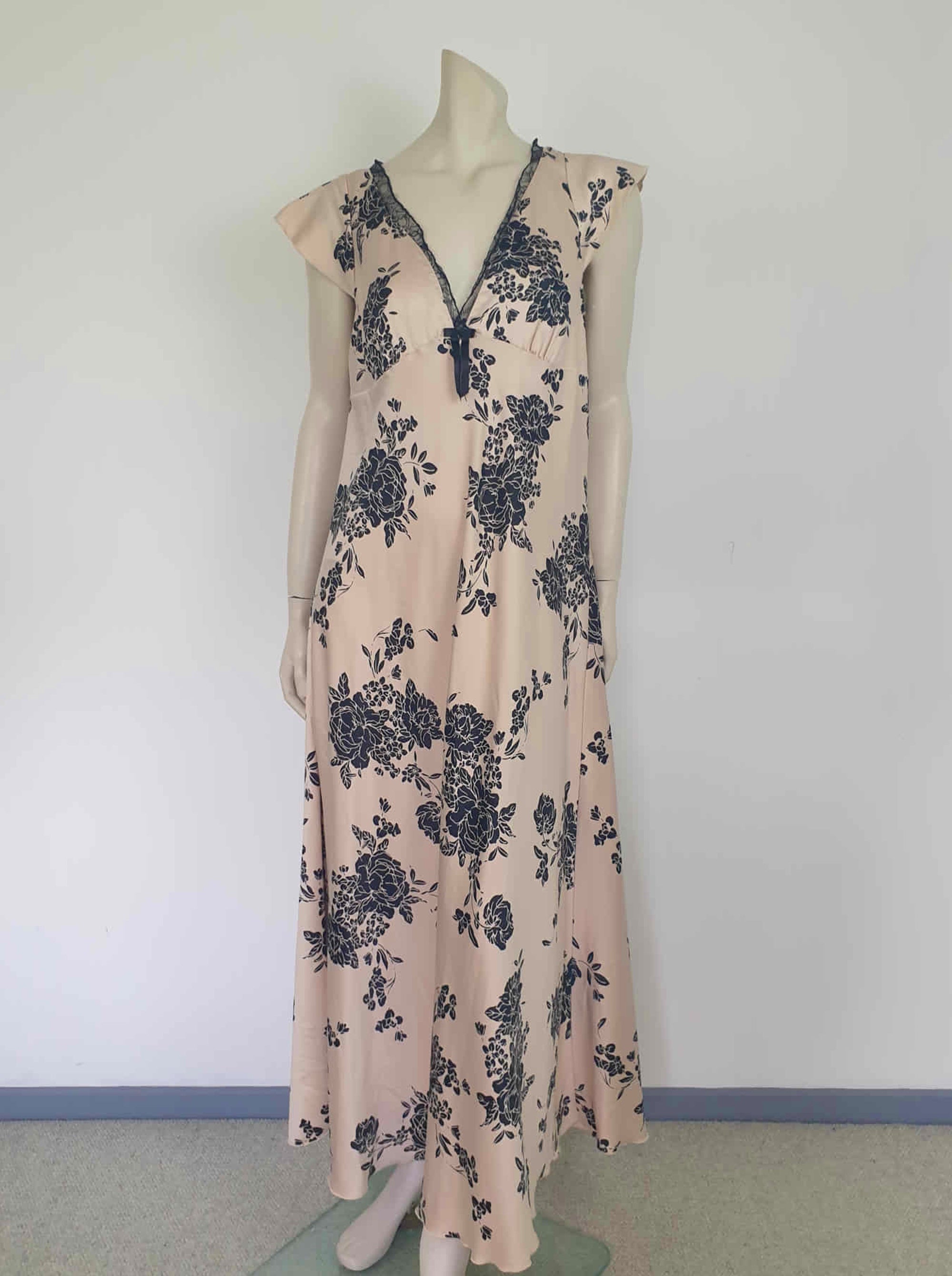 Long beige satin nightgown with large black floral print and flared capped sleeves. The skirt falls from beneath the bust. Slit to the thigh on one side. The V neck is trimmed with narrow black lace and a bow. Elasticised across the back. Fabric is polyester satin. Brand is illegible. 1990s-Y2K?