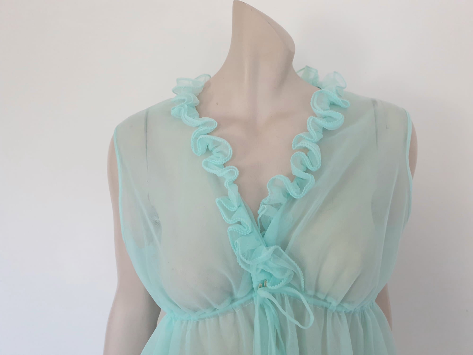Sheer aqua mini nightgown has a crossover V neck with a drawstring beneath at the front. Double ruffle at the neckline. The back is quite full. Mid-thigh length. It's made from two layers of totally sheer nylon. Brand is Linella - 1960s. Medium.