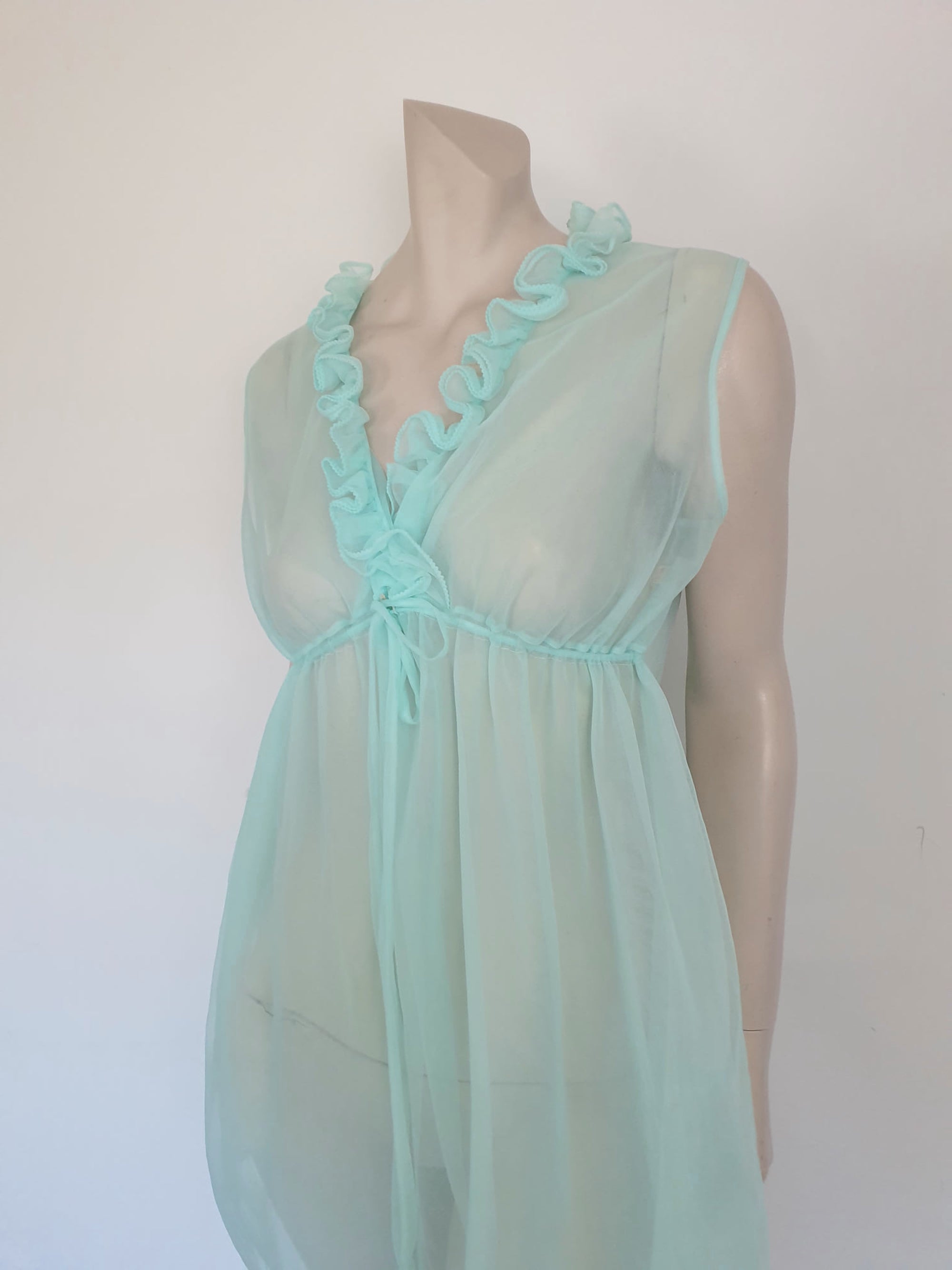 Sheer aqua mini nightgown has a crossover V neck with a drawstring beneath at the front. Double ruffle at the neckline. The back is quite full. Mid-thigh length. It's made from two layers of totally sheer nylon. Brand is Linella - 1960s. Medium.