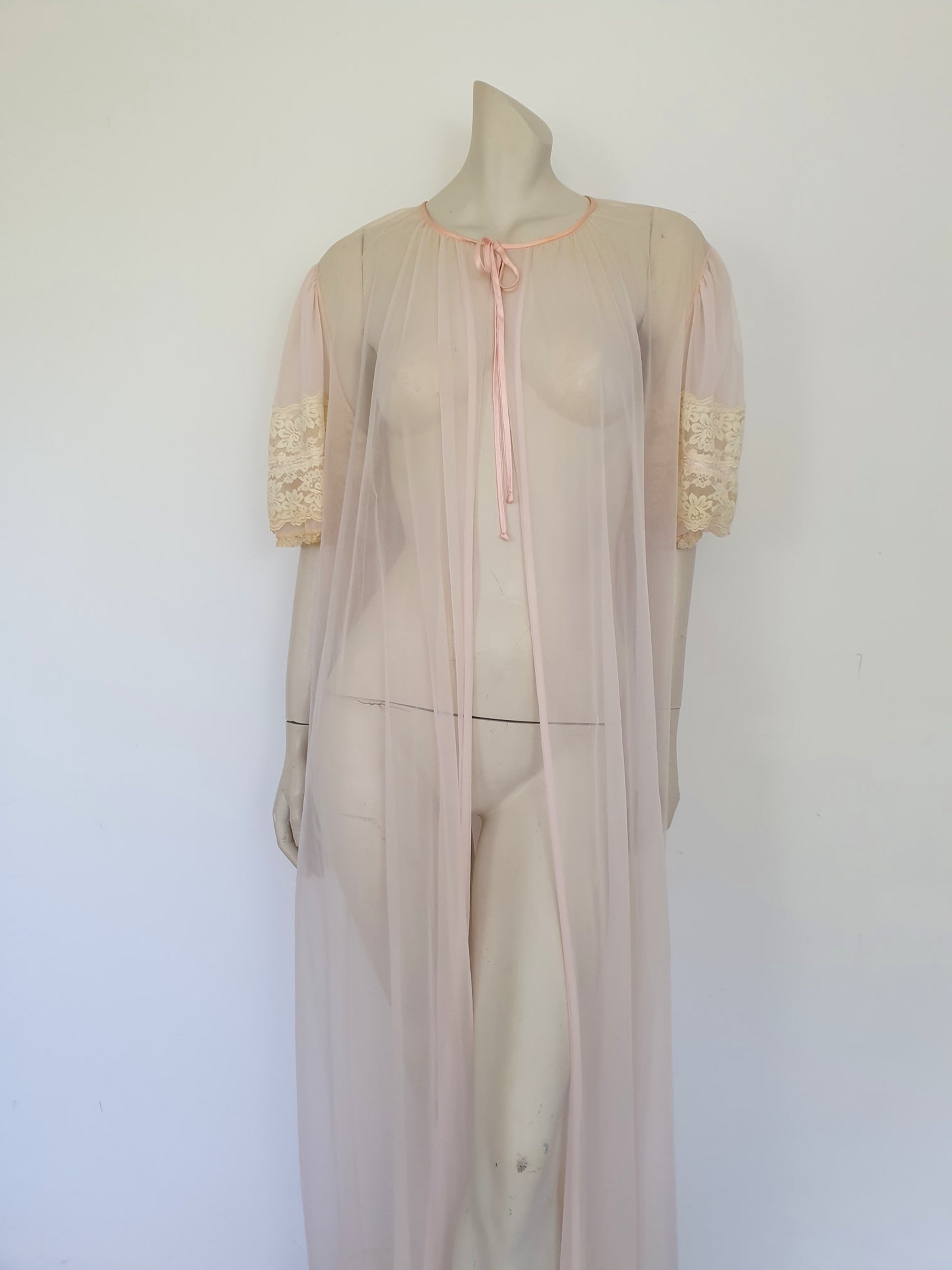 Sheer peach peignoir or long robe has puff sleeves trimmed with wide lace. The hem is also trimmed with wide ecru (beige) lace. The robe is gathered into the neckline, which is bound with satin and extends into a tie at the front. Fabric is a single layer of sheerest nylon. The tag is illegible - 1960s. Large.