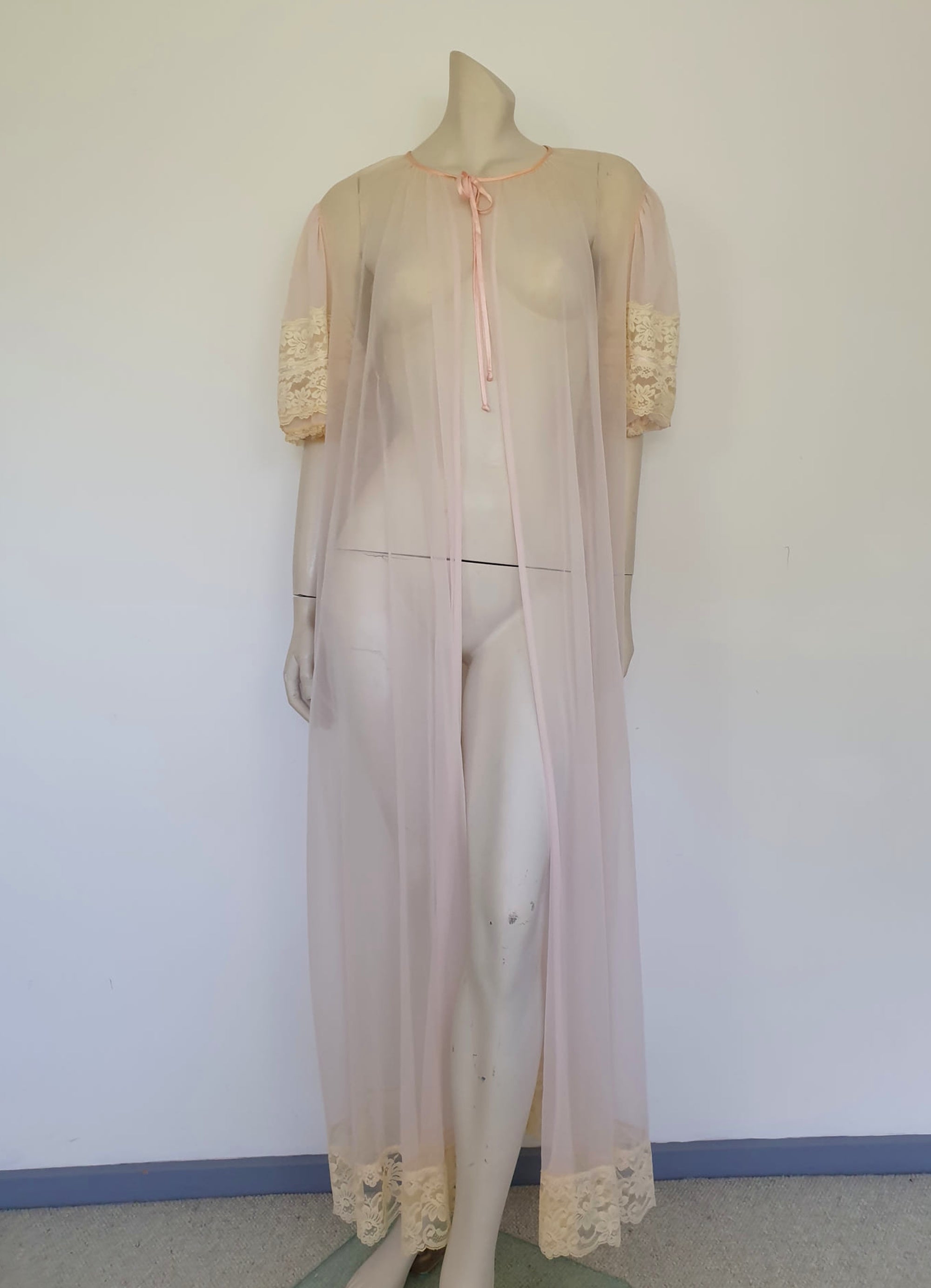 Sheer peach peignoir or long robe has puff sleeves trimmed with wide lace. The hem is also trimmed with wide ecru (beige) lace. The robe is gathered into the neckline, which is bound with satin and extends into a tie at the front. Fabric is a single layer of sheerest nylon. The tag is illegible - 1960s. Large.