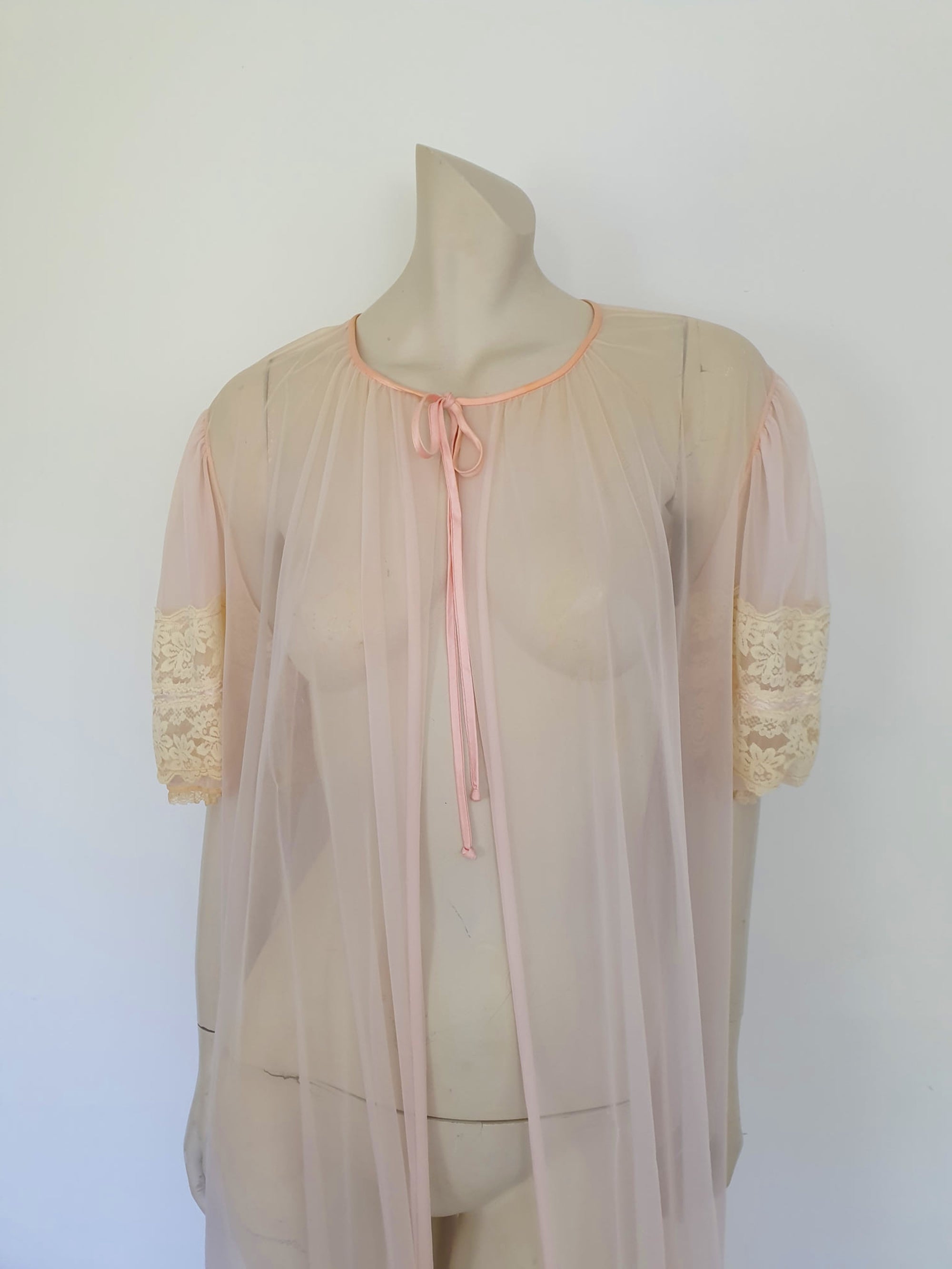 Sheer peach peignoir or long robe has puff sleeves trimmed with wide lace. The hem is also trimmed with wide ecru (beige) lace. The robe is gathered into the neckline, which is bound with satin and extends into a tie at the front. Fabric is a single layer of sheerest nylon. The tag is illegible - 1960s. Large.