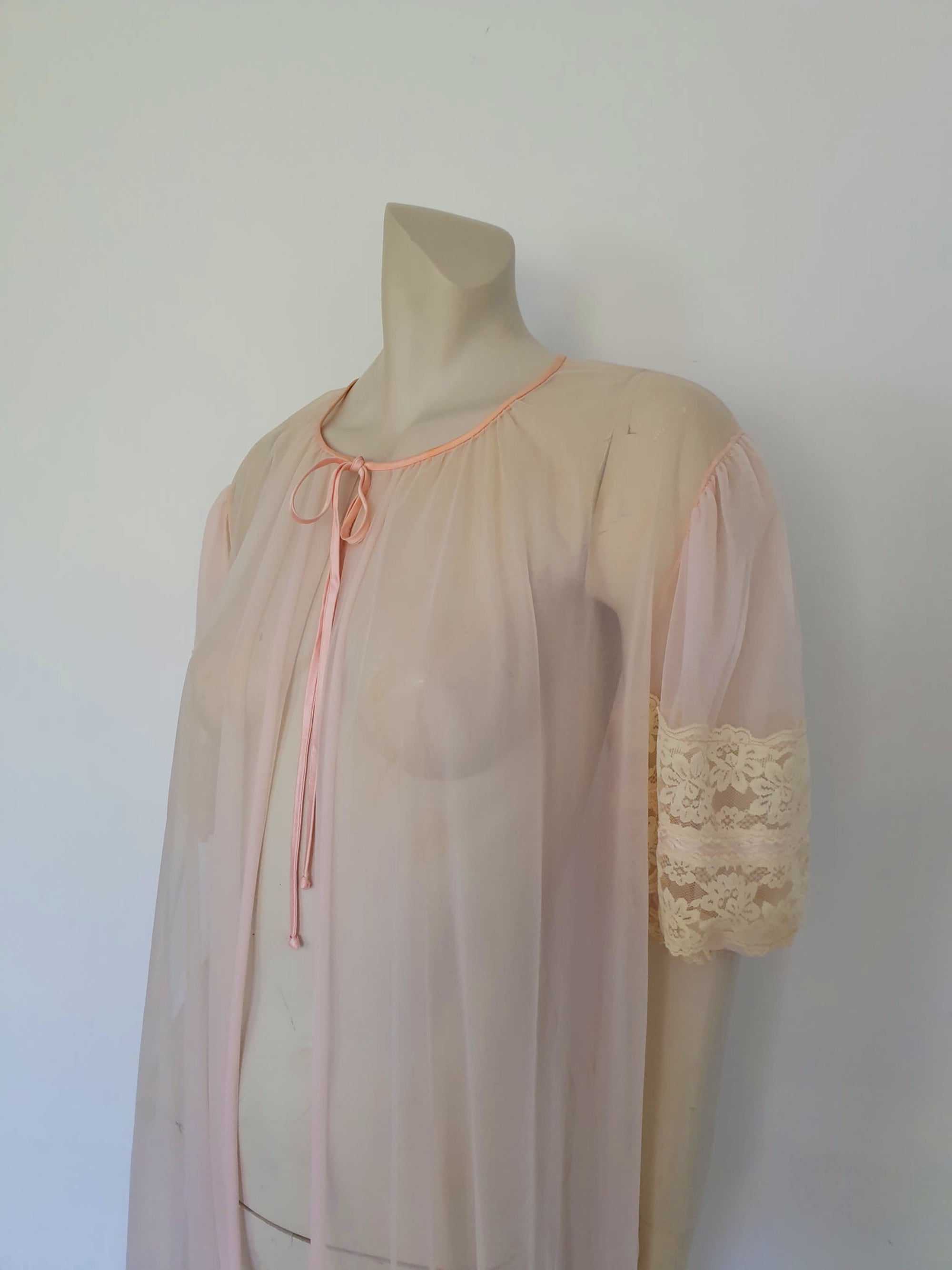 Sheer peach peignoir or long robe has puff sleeves trimmed with wide lace. The hem is also trimmed with wide ecru (beige) lace. The robe is gathered into the neckline, which is bound with satin and extends into a tie at the front. Fabric is a single layer of sheerest nylon. The tag is illegible - 1960s. Large.