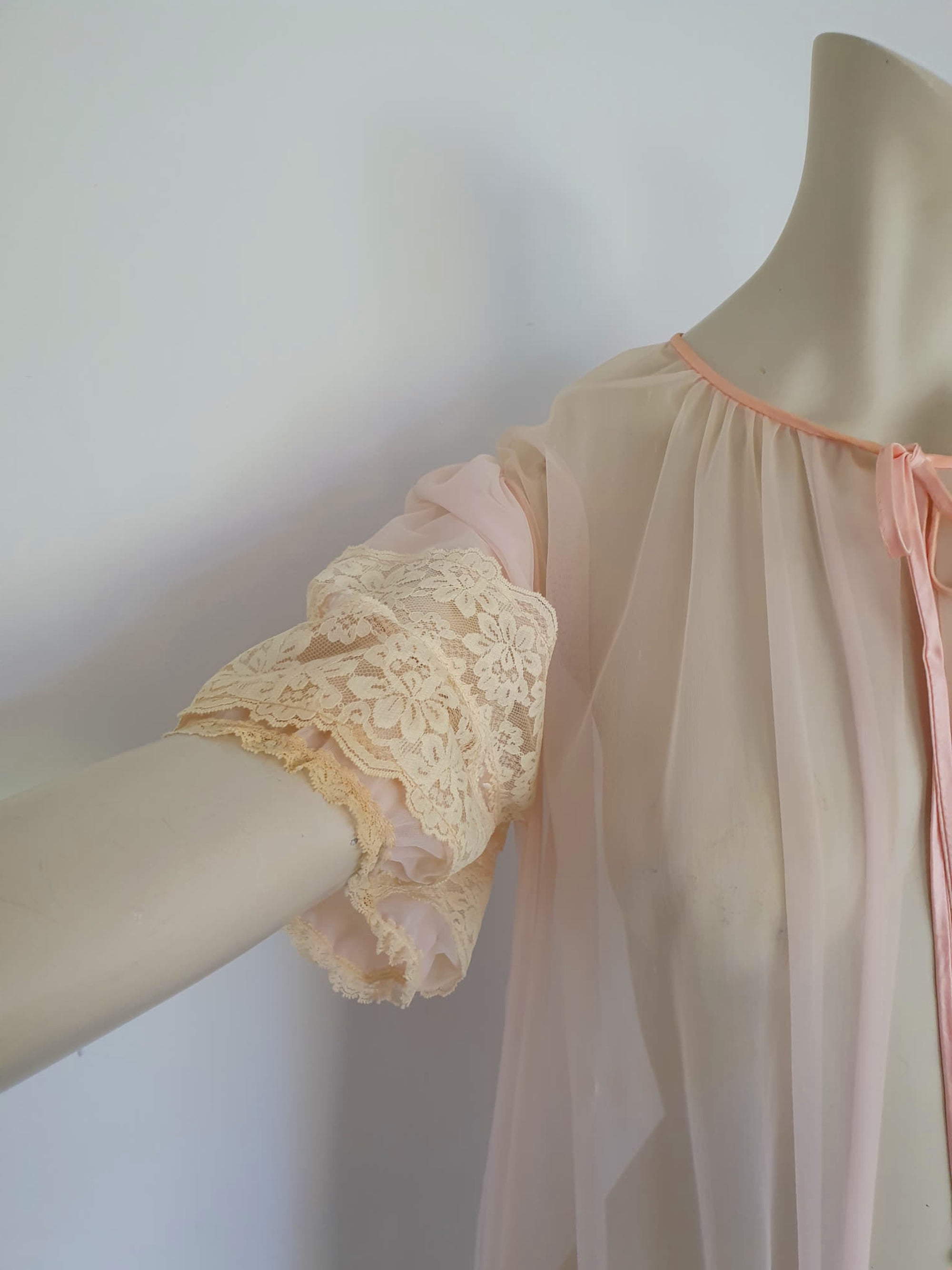 Sheer peach peignoir or long robe has puff sleeves trimmed with wide lace. The hem is also trimmed with wide ecru (beige) lace. The robe is gathered into the neckline, which is bound with satin and extends into a tie at the front. Fabric is a single layer of sheerest nylon. The tag is illegible - 1960s. Large.