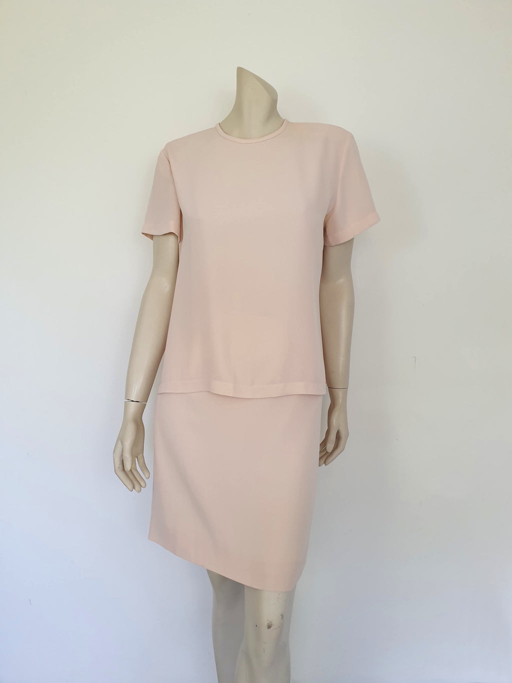 Above-knee length pale pink skirt and top set. Boxy top has short sleeves and is a pullover style, with a short opening buttoning at the back of the neck. There are removable shoulder pads. The straight skirt has a rear vent, rear zip and button fastening and is lined. Fabric is polyester crepe. Made in New Zealand for Suzanne Gregory - 1980s. Medium to Large.