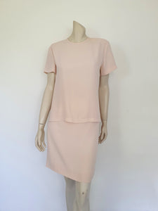 Above-knee length pale pink skirt and top set. Boxy top has short sleeves and is a pullover style, with a short opening buttoning at the back of the neck. There are removable shoulder pads. The straight skirt has a rear vent, rear zip and button fastening and is lined. Fabric is polyester crepe. Made in New Zealand for Suzanne Gregory - 1980s. Medium to Large.