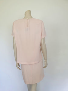Above-knee length pale pink skirt and top set. Boxy top has short sleeves and is a pullover style, with a short opening buttoning at the back of the neck. There are removable shoulder pads. The straight skirt has a rear vent, rear zip and button fastening and is lined. Fabric is polyester crepe. Made in New Zealand for Suzanne Gregory - 1980s. Medium to Large.