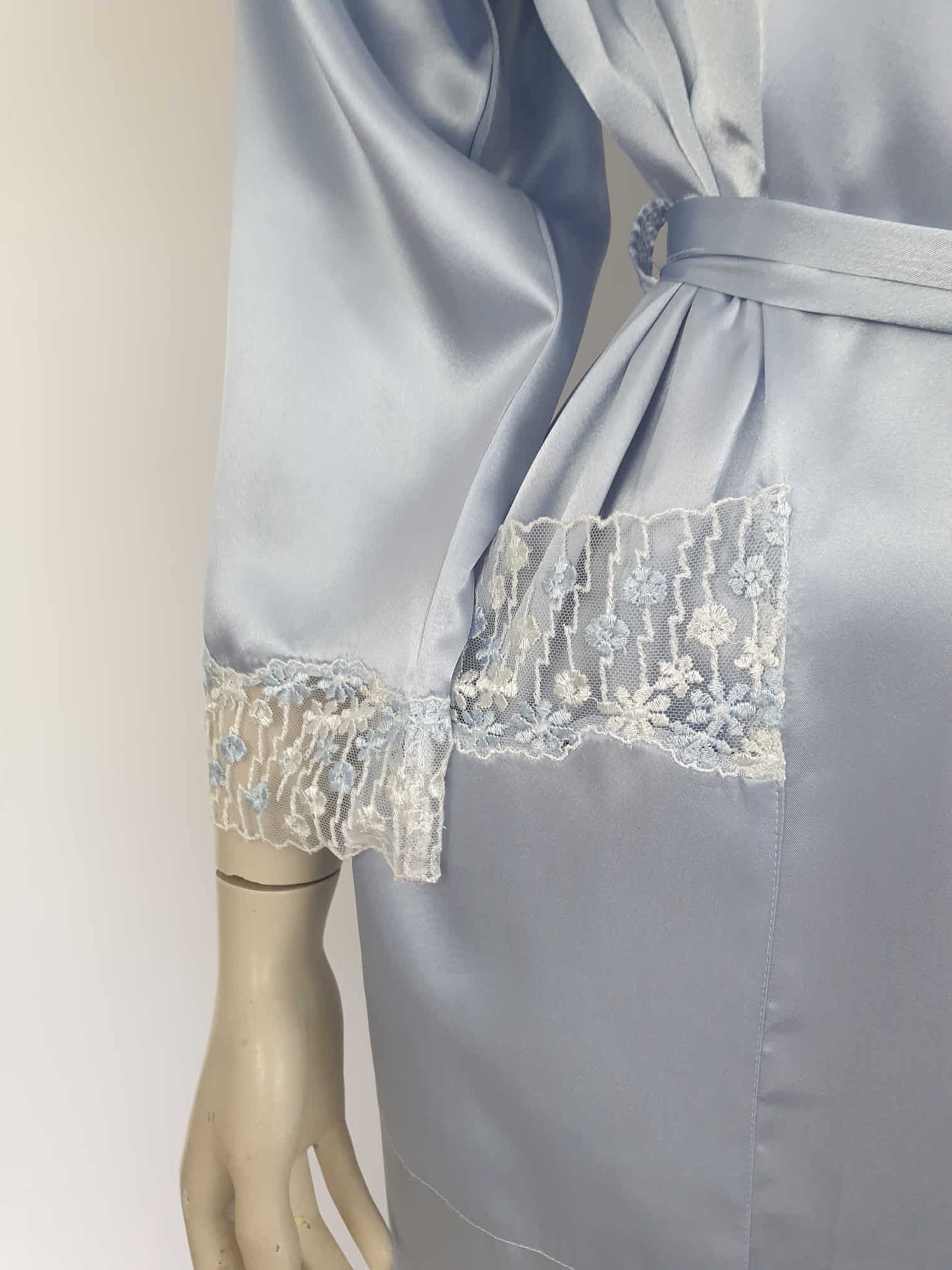 Beautiful blue satin robe with two patch pockets. Sleeves and pockets are trimmed with wide blue and white lace. Shawl collar and separate tie through carriers at the waist. Shoulder pads. Fabric is polyester satin. Made under licence in Australia for a famous French designer, see label - 1980s.