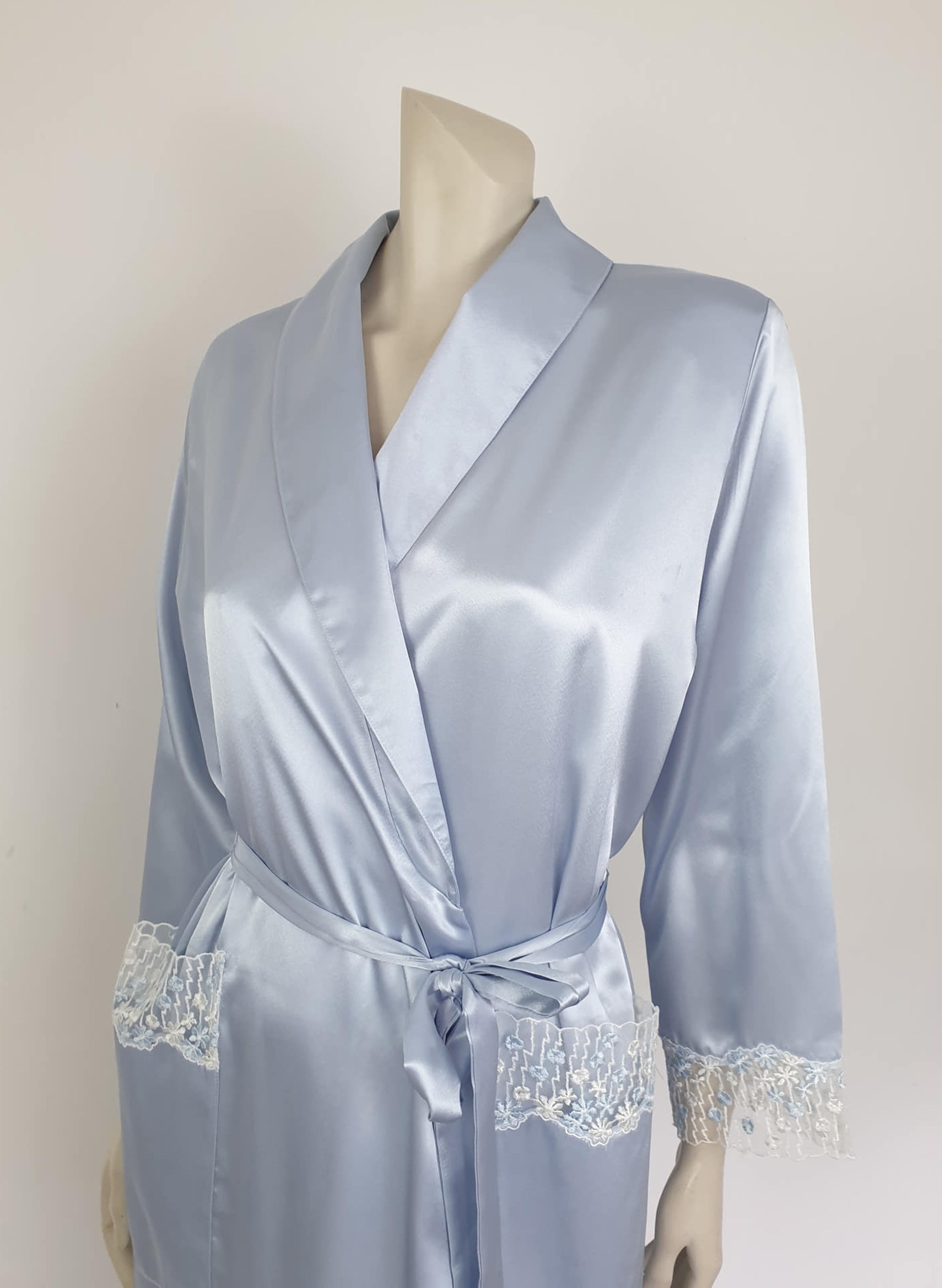 Beautiful blue satin robe with two patch pockets. Sleeves and pockets are trimmed with wide blue and white lace. Shawl collar and separate tie through carriers at the waist. Shoulder pads. Fabric is polyester satin. Made under licence in Australia for a famous French designer, see label - 1980s.