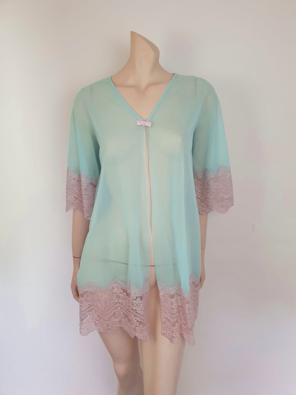 Thigh length robe has a V-neckline and wide pinky-beige lace at the below-elbow length sleeves and hem. Small bow at the neck. Closes at the front with one press stud.  The fabric is a double layer of sheer nylon. Made in the USA for Gilead - 1960s. Medium