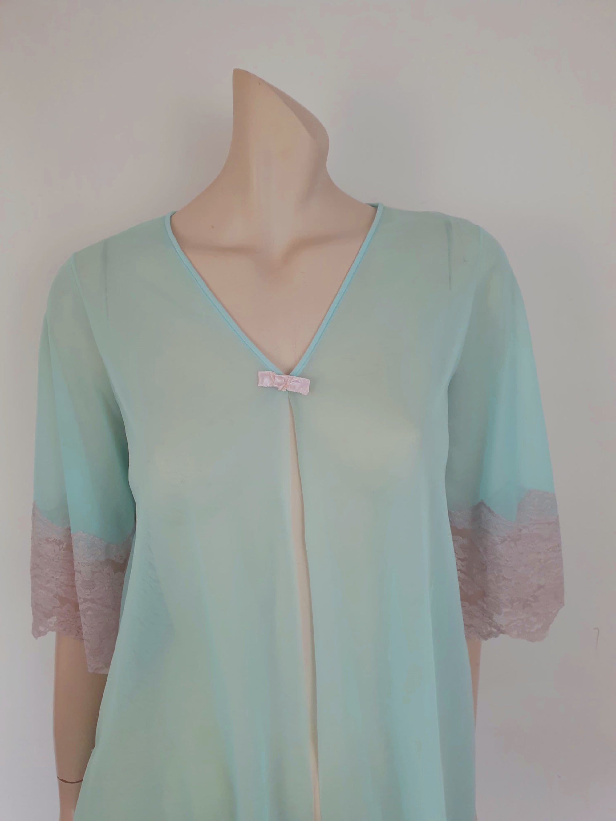 Thigh length robe has a V-neckline and wide pinky-beige lace at the below-elbow length sleeves and hem. Small bow at the neck. Closes at the front with one press stud.  The fabric is a double layer of sheer nylon. Made in the USA for Gilead - 1960s. Medium