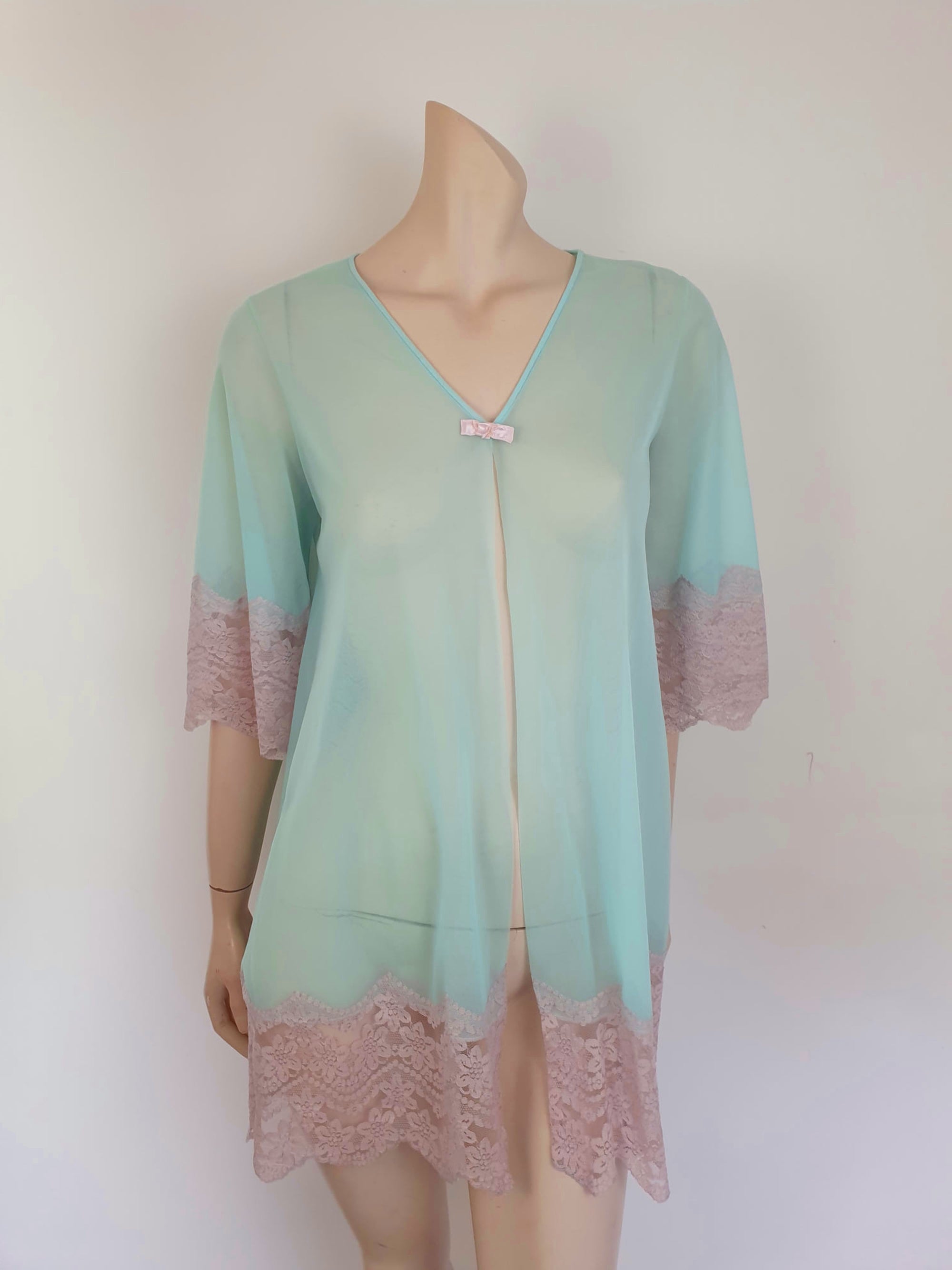 Thigh length robe has a V-neckline and wide pinky-beige lace at the below-elbow length sleeves and hem. Small bow at the neck. Closes at the front with one press stud.  The fabric is a double layer of sheer nylon. Made in the USA for Gilead - 1960s. Medium