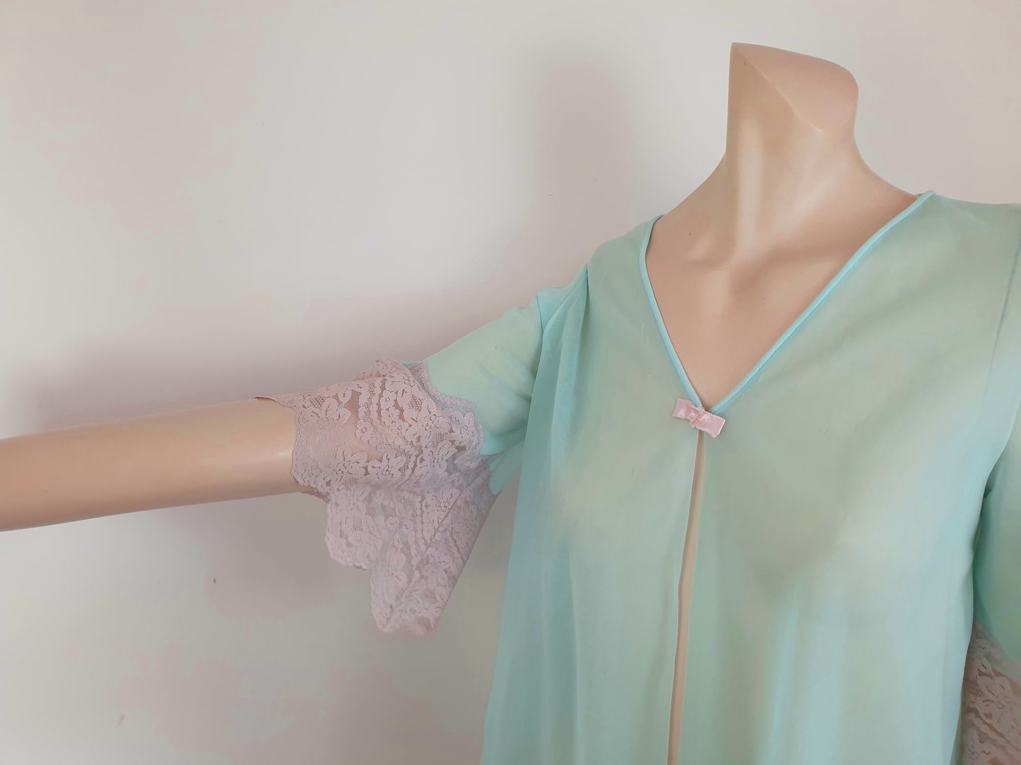 Thigh length robe has a V-neckline and wide pinky-beige lace at the below-elbow length sleeves and hem. Small bow at the neck. Closes at the front with one press stud.  The fabric is a double layer of sheer nylon. Made in the USA for Gilead - 1960s. Medium
