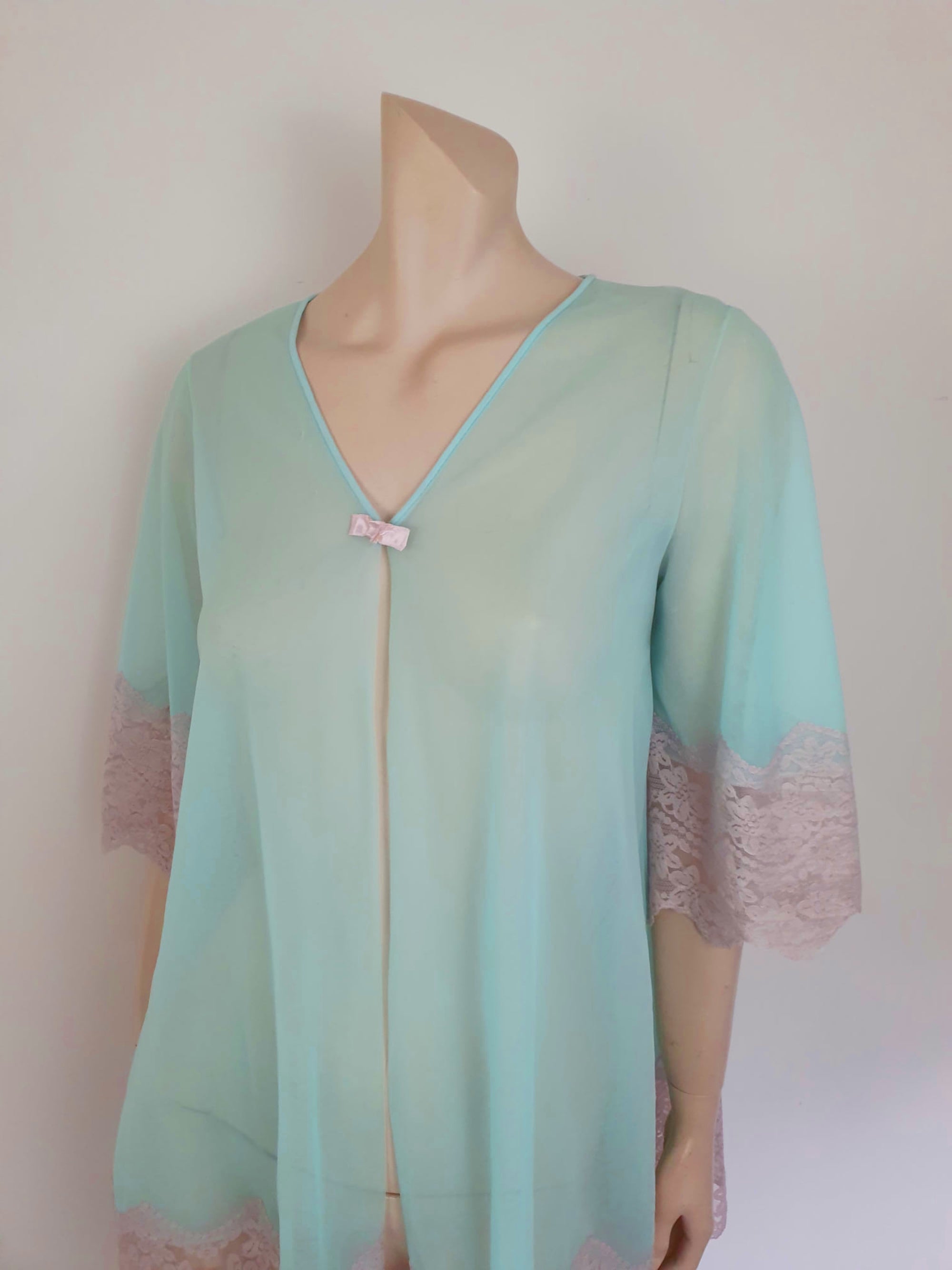 Thigh length robe has a V-neckline and wide pinky-beige lace at the below-elbow length sleeves and hem. Small bow at the neck. Closes at the front with one press stud.  The fabric is a double layer of sheer nylon. Made in the USA for Gilead - 1960s. Medium