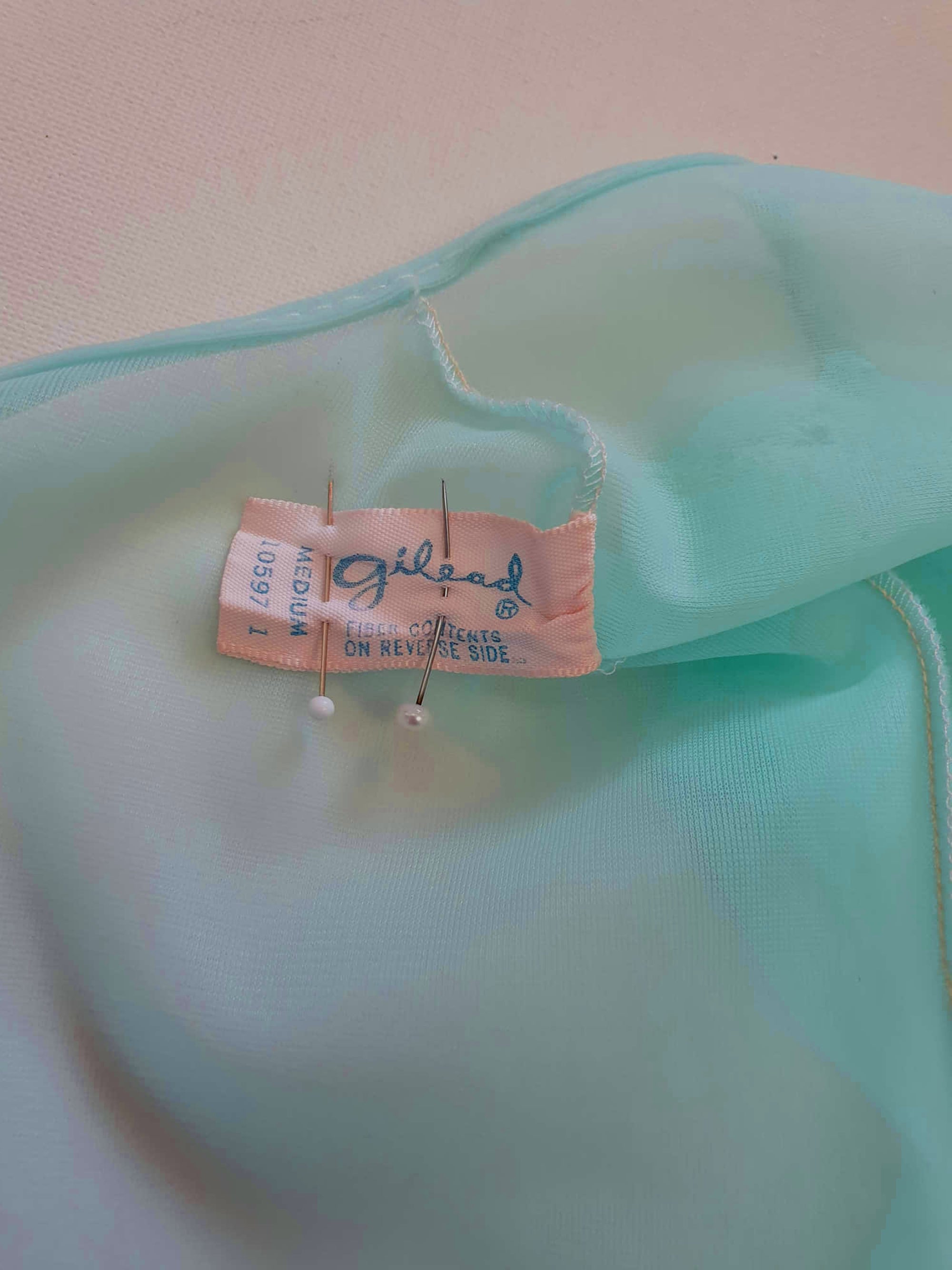 Thigh length robe has a V-neckline and wide pinky-beige lace at the below-elbow length sleeves and hem. Small bow at the neck. Closes at the front with one press stud.  The fabric is a double layer of sheer nylon. Made in the USA for Gilead - 1960s. Medium