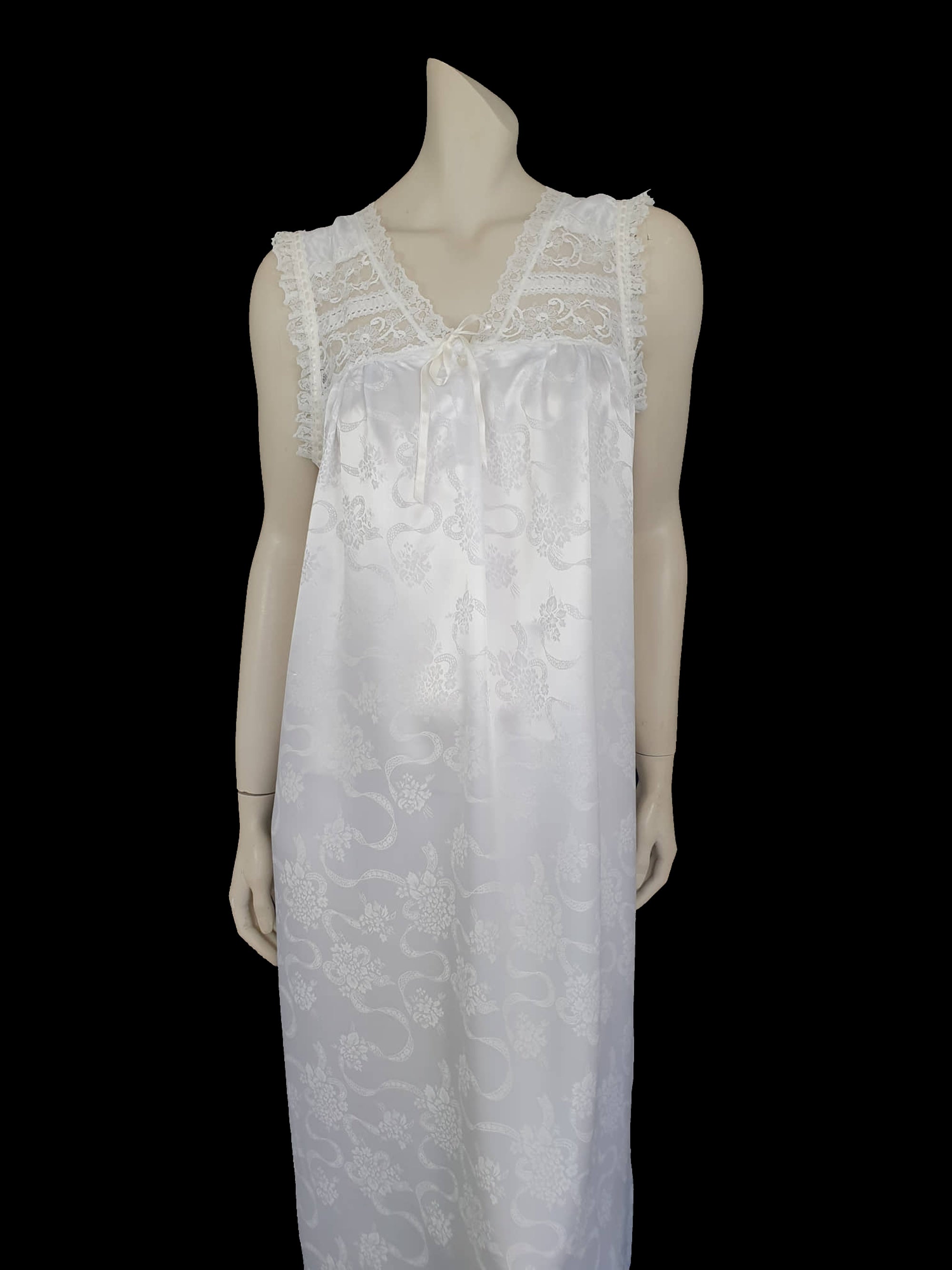 White satin nightgown has a lacy yoke, V-neck and has lace trim at the neckline, armholes and hem. The beautiful fabric is a damask weave with a floral design. Closes at the neck with one button and a ribbon bow. The nightie is mid to lower calf length. Fabric is polyester. Made in Australia by Texwear - 1980s. Medium