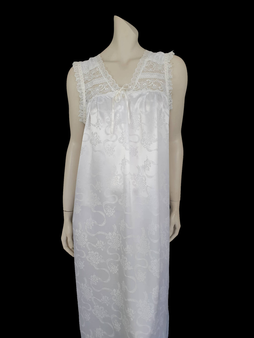 White satin nightgown has a lacy yoke, V-neck and has lace trim at the neckline, armholes and hem. The beautiful fabric is a damask weave with a floral design. Closes at the neck with one button and a ribbon bow. The nightie is mid to lower calf length. Fabric is polyester. Made in Australia by Texwear - 1980s. Medium