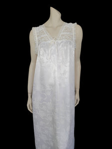 White satin nightgown has a lacy yoke, V-neck and has lace trim at the neckline, armholes and hem. The beautiful fabric is a damask weave with a floral design. Closes at the neck with one button and a ribbon bow. The nightie is mid to lower calf length. Fabric is polyester. Made in Australia by Texwear - 1980s. Medium